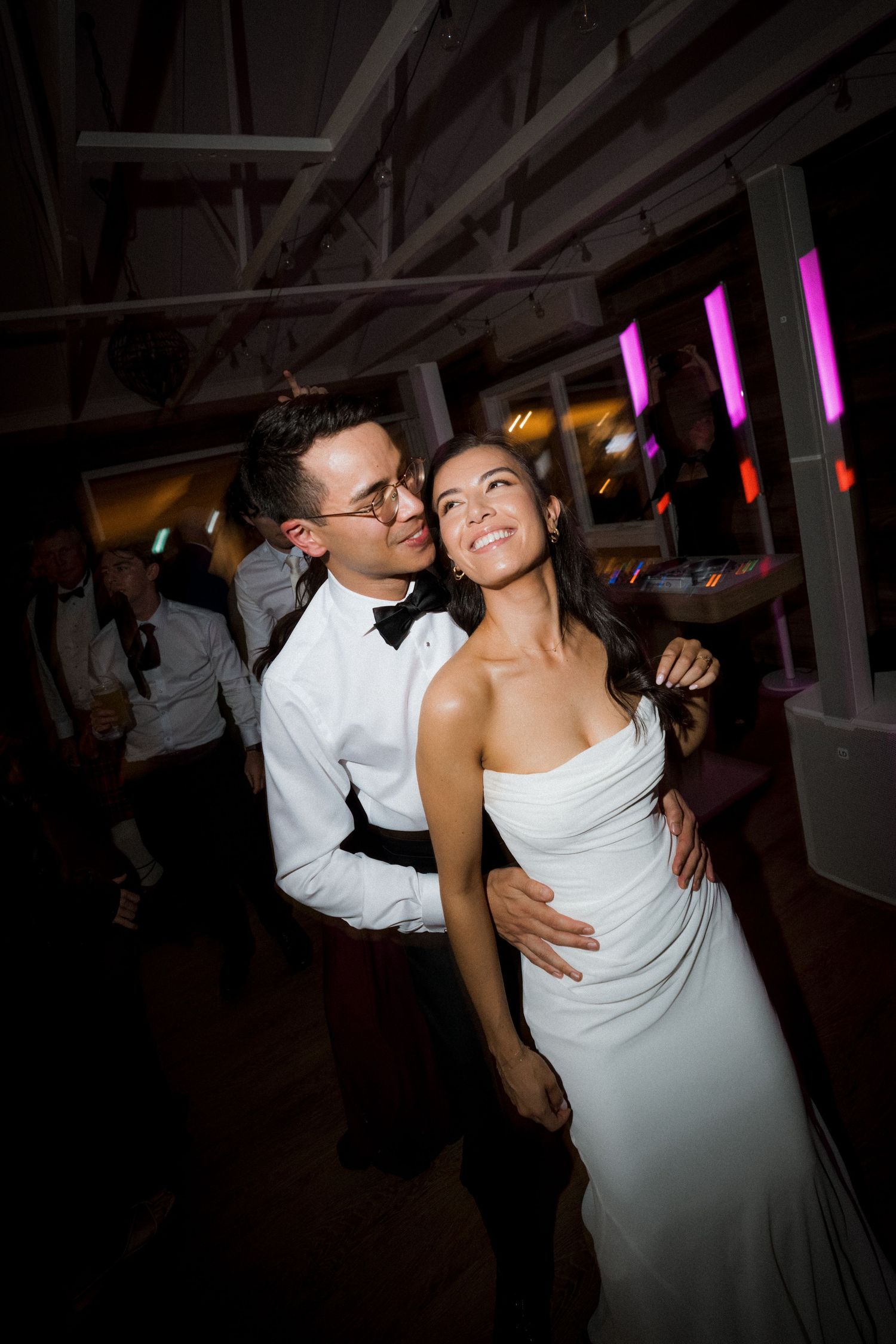 Groom in white shirt and bow tie embraces smiling bride during wedding reception dance.