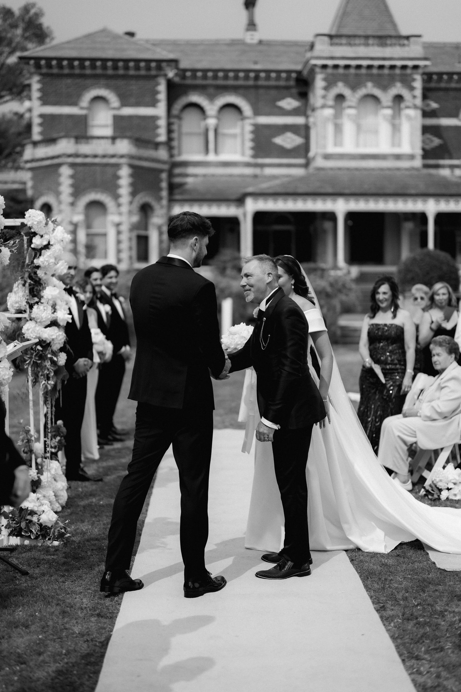 An outdoor wedding ceremony takes place in front of a historic Victorian mansion with guests watching from white chairs.