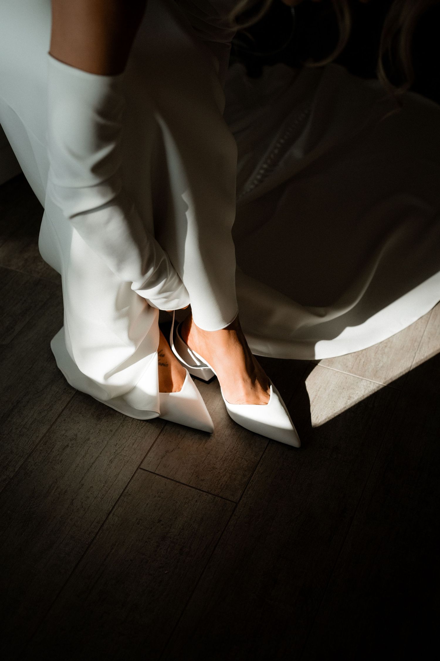 Elegant white bridal shoe highlighted by dramatic lighting on a dark wooden floor.