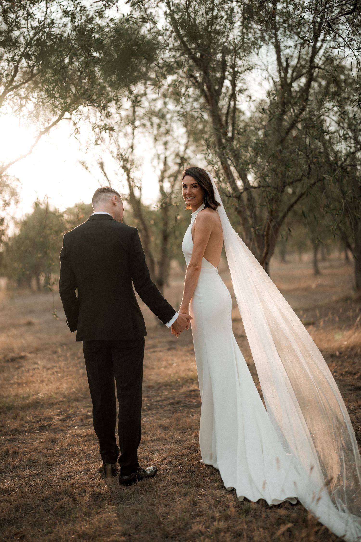 A bride in a flowing white gown looks back while holding hands with groom during sunset outdoor photo session.