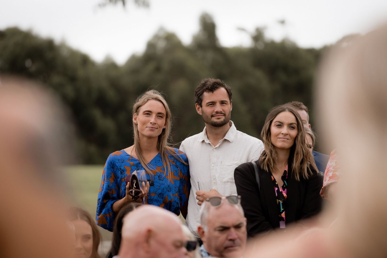 Three people in casual attire standing together outdoors with blurred figures in the foreground.