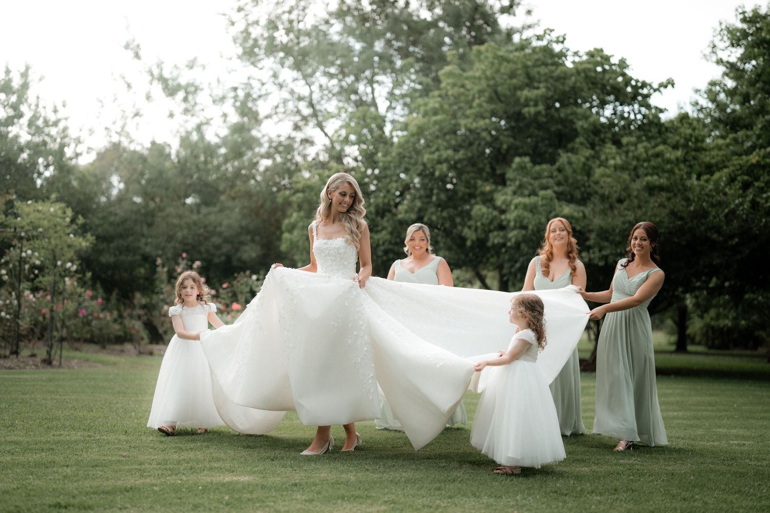 Playful wedding party photo with flower girls helping to spread the bride's dress in a garden.