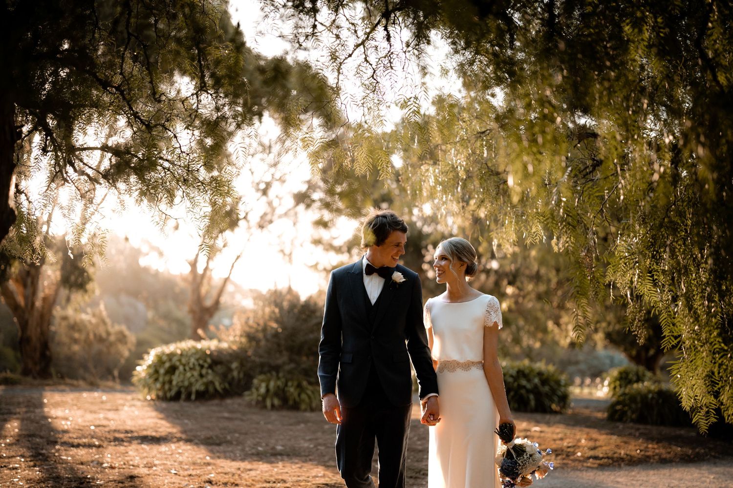 A romantic golden hour wedding photo of newlyweds walking together under willow trees along a garden path.