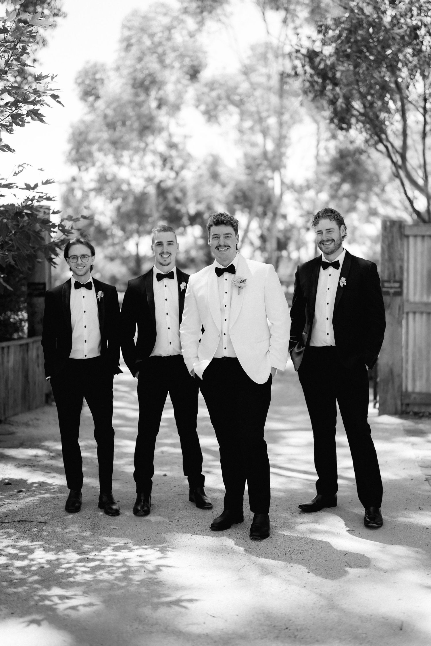 Groom in white tuxedo jacket poses with three groomsmen in black tuxedos in black and white wedding photo.