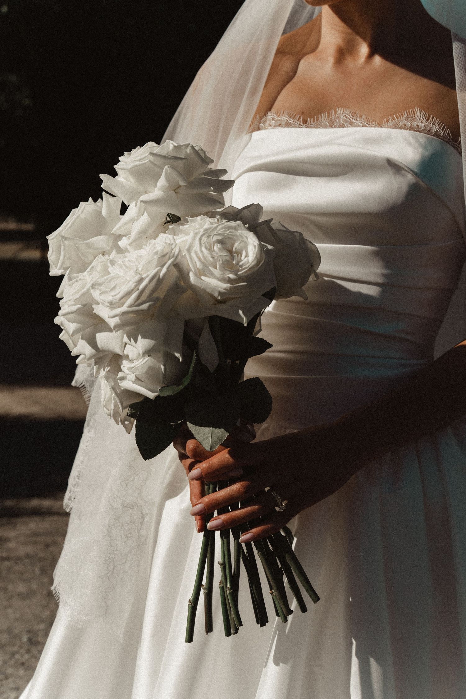 Bride in white off-shoulder gown holding elegant bouquet of white roses with dark green stems in soft sunlight.