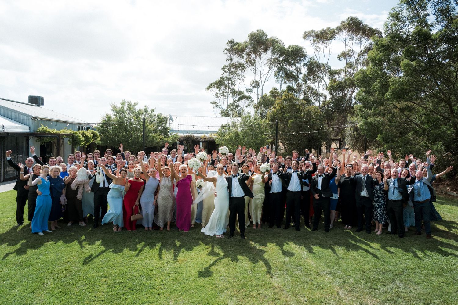Large wedding party group photo with guests gathered outdoors on a grassy hillside.