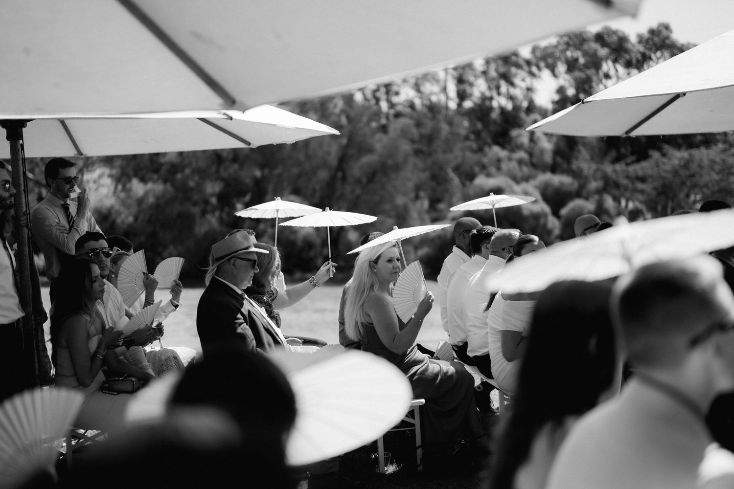 Wedding guests hold white parasols during an outdoor ceremony.