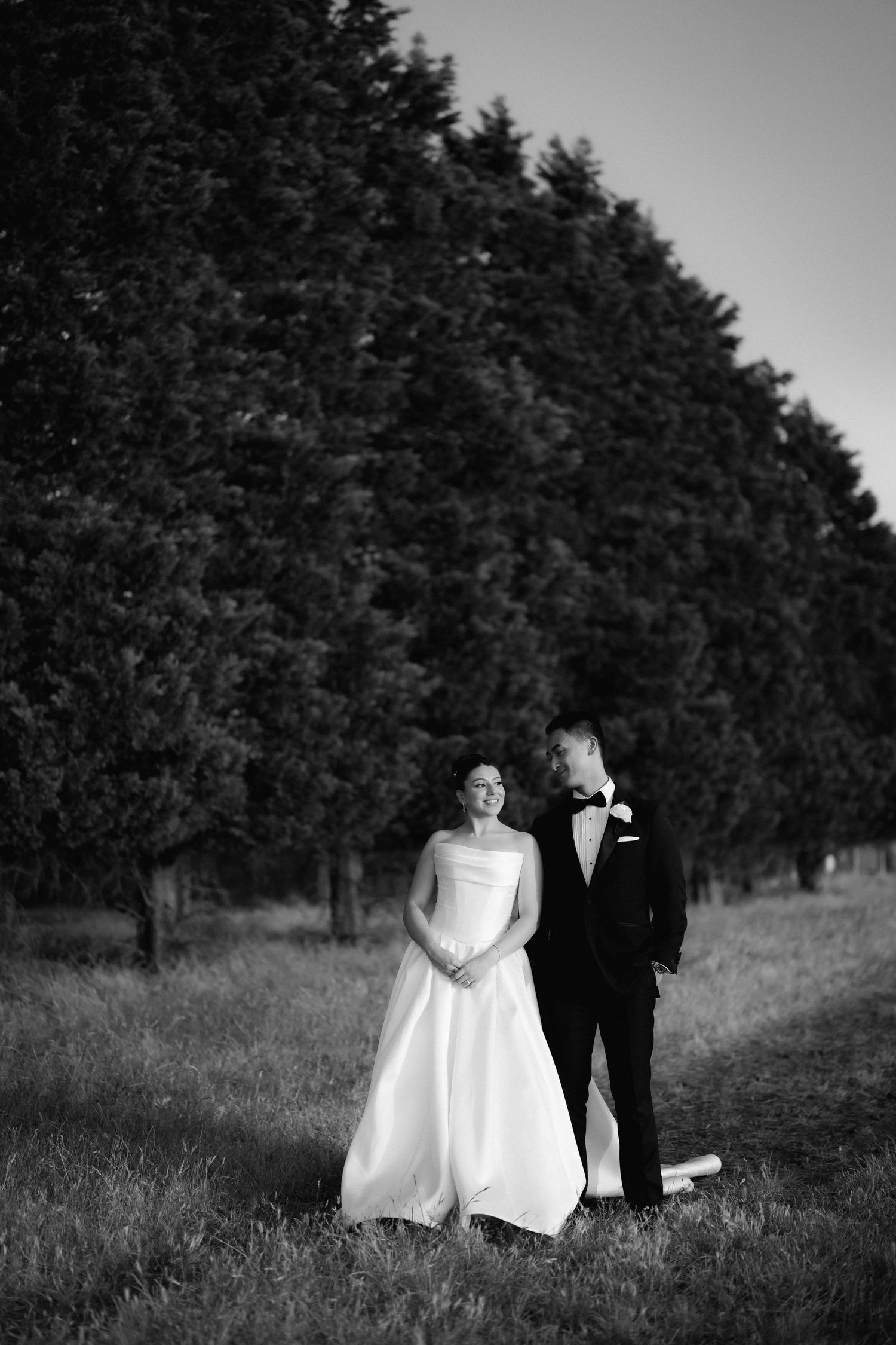 Black and white photo of a couple walking along a tree-lined path in formal attire.