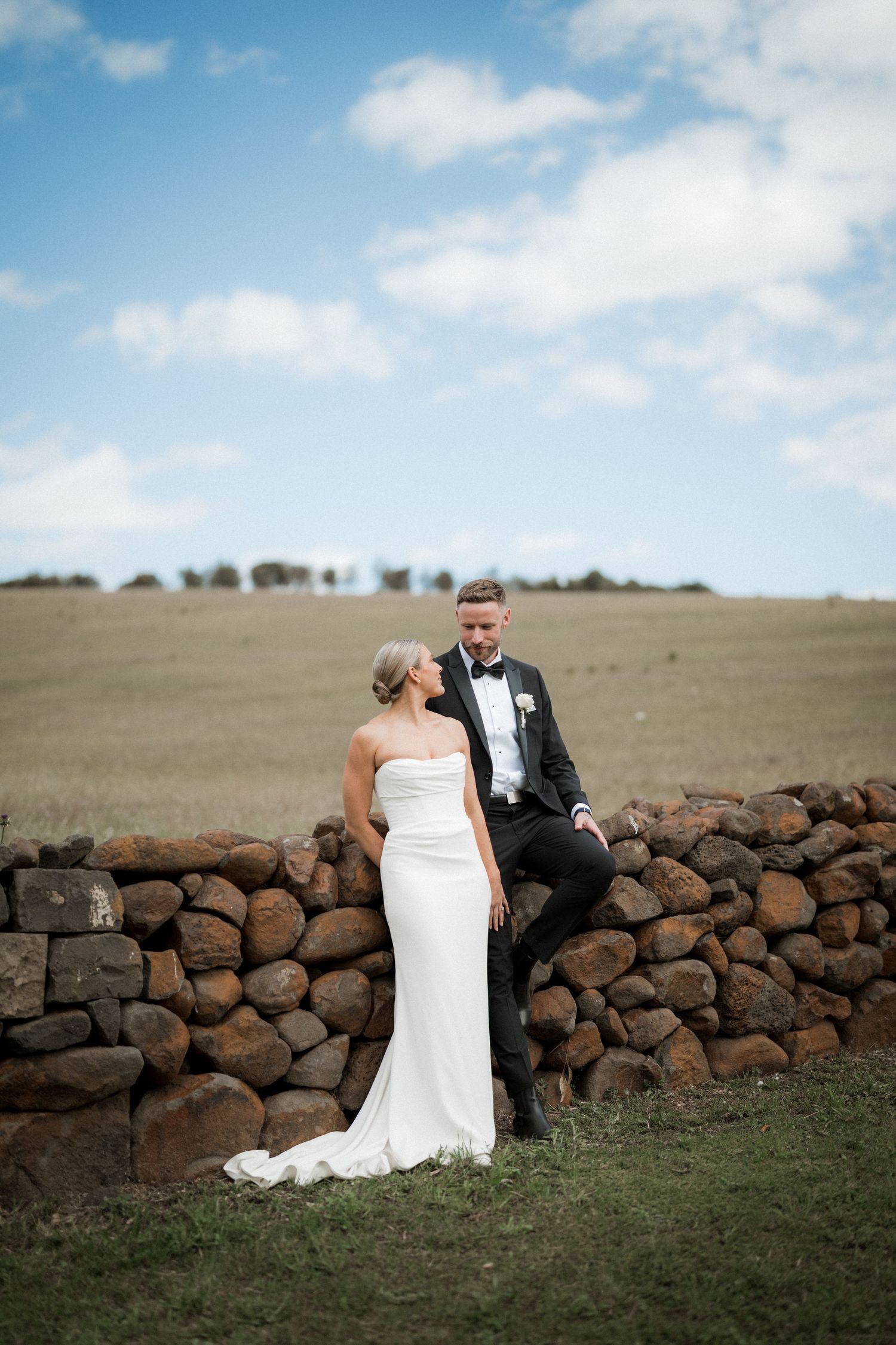 Wedding couple posing by rustic stone wall in countryside setting.