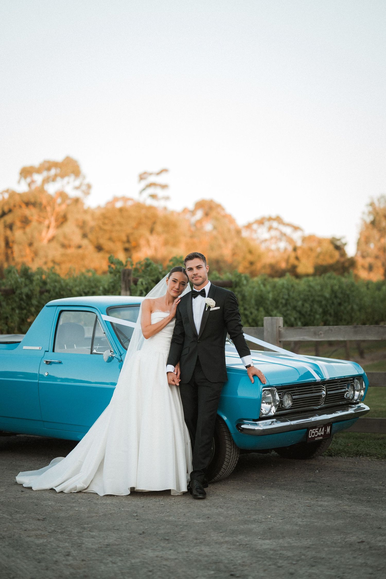 Wedding couple poses with vintage blue car during golden hour photography session.