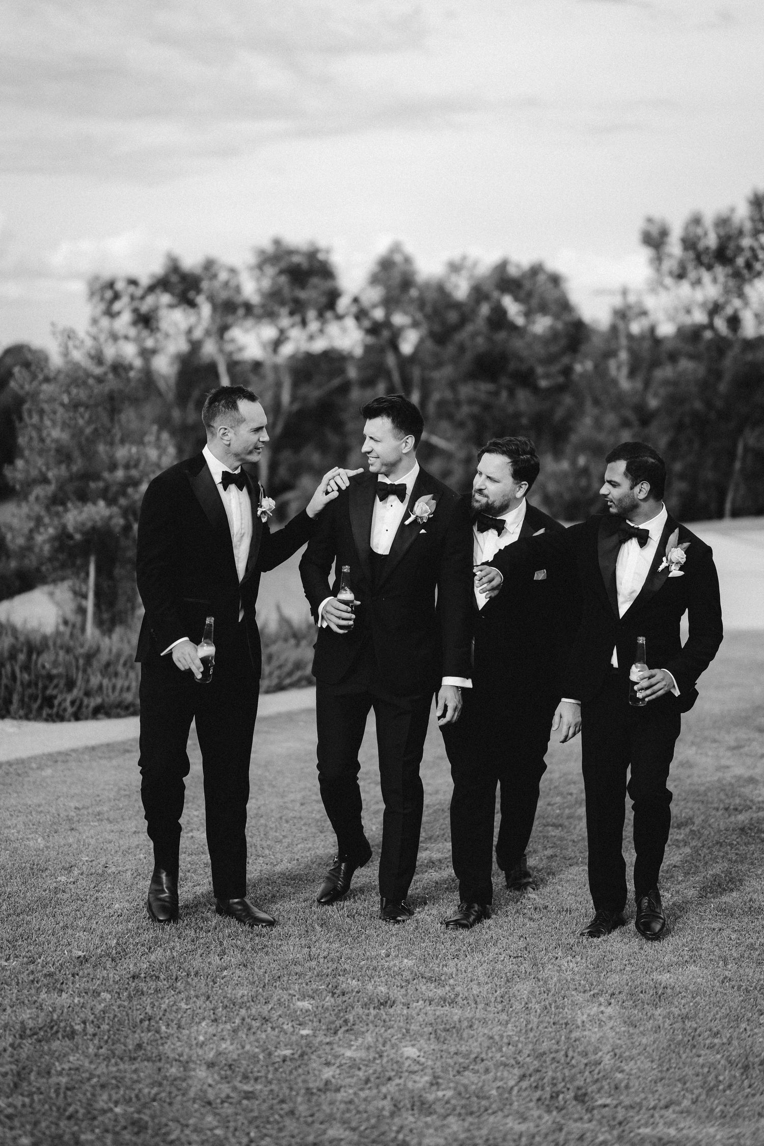 Four groomsmen in black tuxedos share a candid laughing moment together on a grassy outdoor venue path.