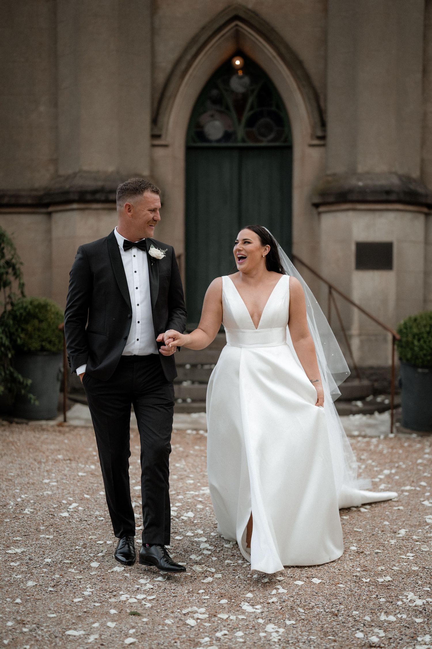 A bride and groom share a joyful moment together while walking outside a gothic church entrance.
