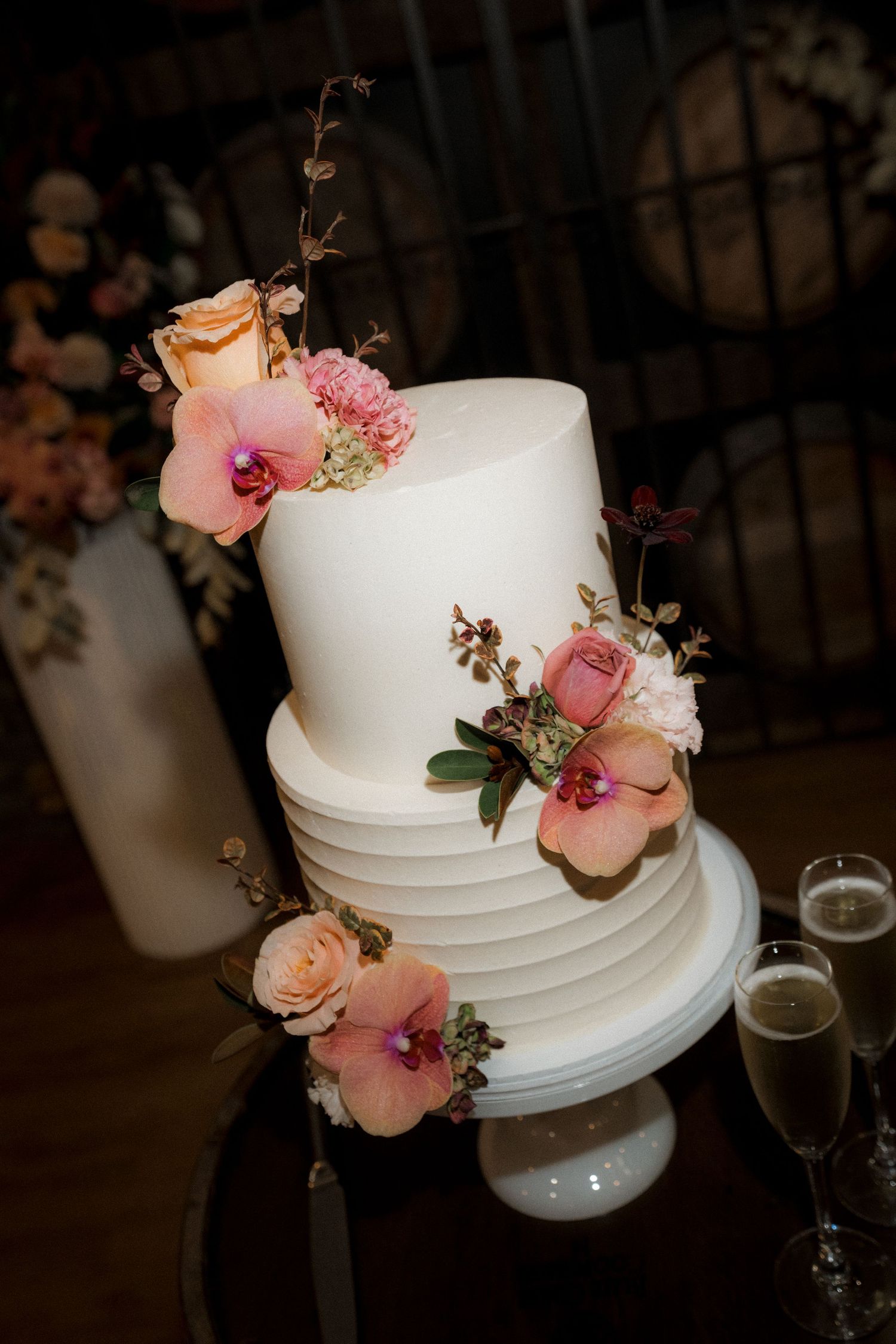 Elegant three-tier white wedding cake adorned with pink orchids and roses on a dark table.
