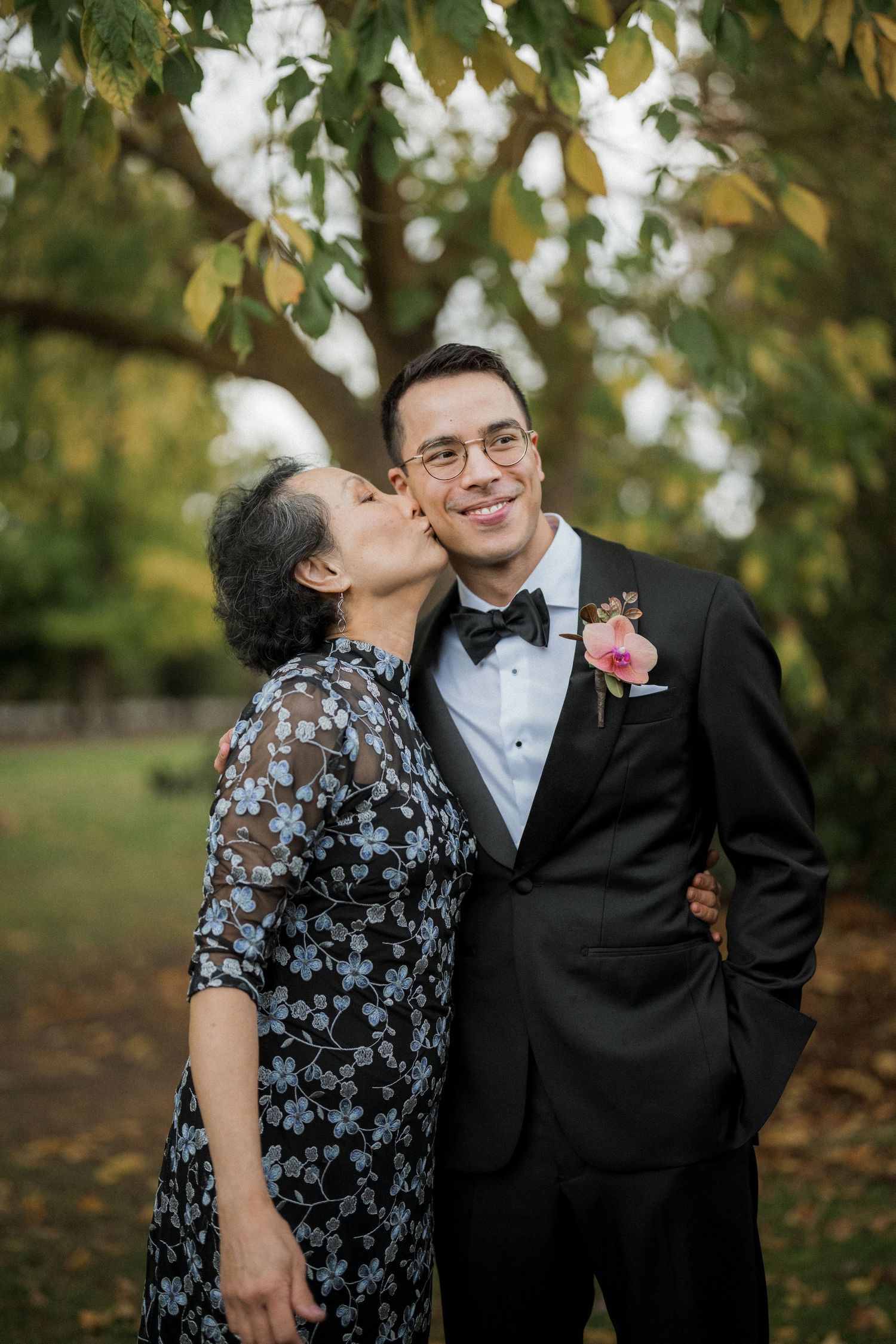 Proud mother kisses her son the groom in a black tuxedo with floral boutonniere outdoors.