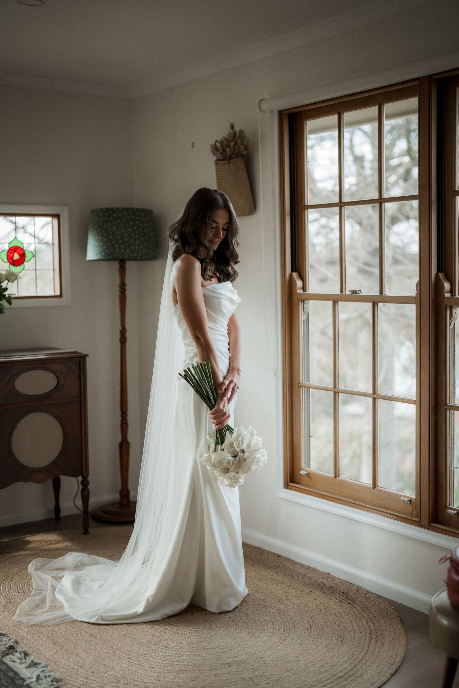 Elegant white wedding dress with flowing train photographed in a sunlit room with wooden windows.