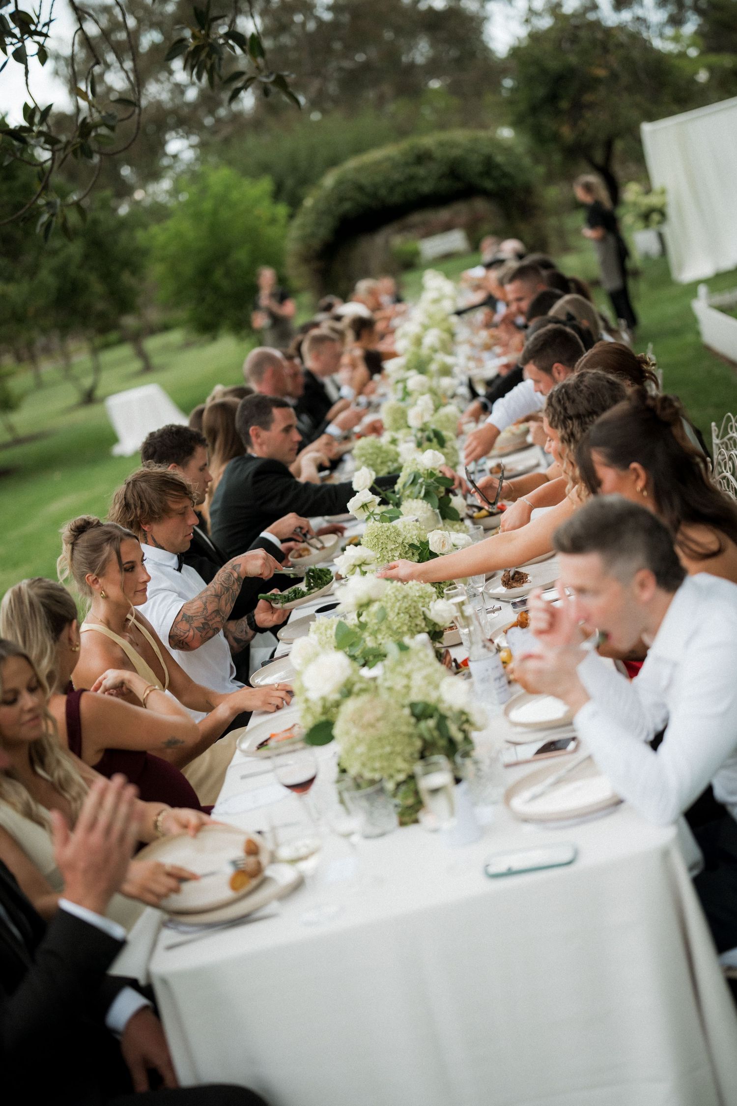 Long wedding reception table with white floral centerpieces and guests dining outdoors.