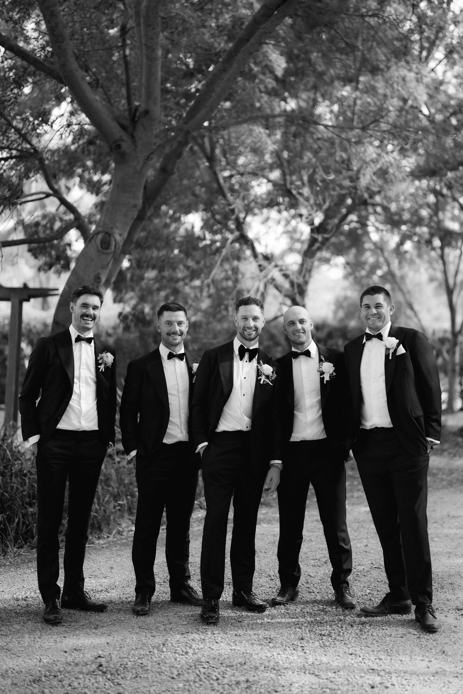 A black and white photo of a wedding party in tuxedos standing together under trees outdoors.