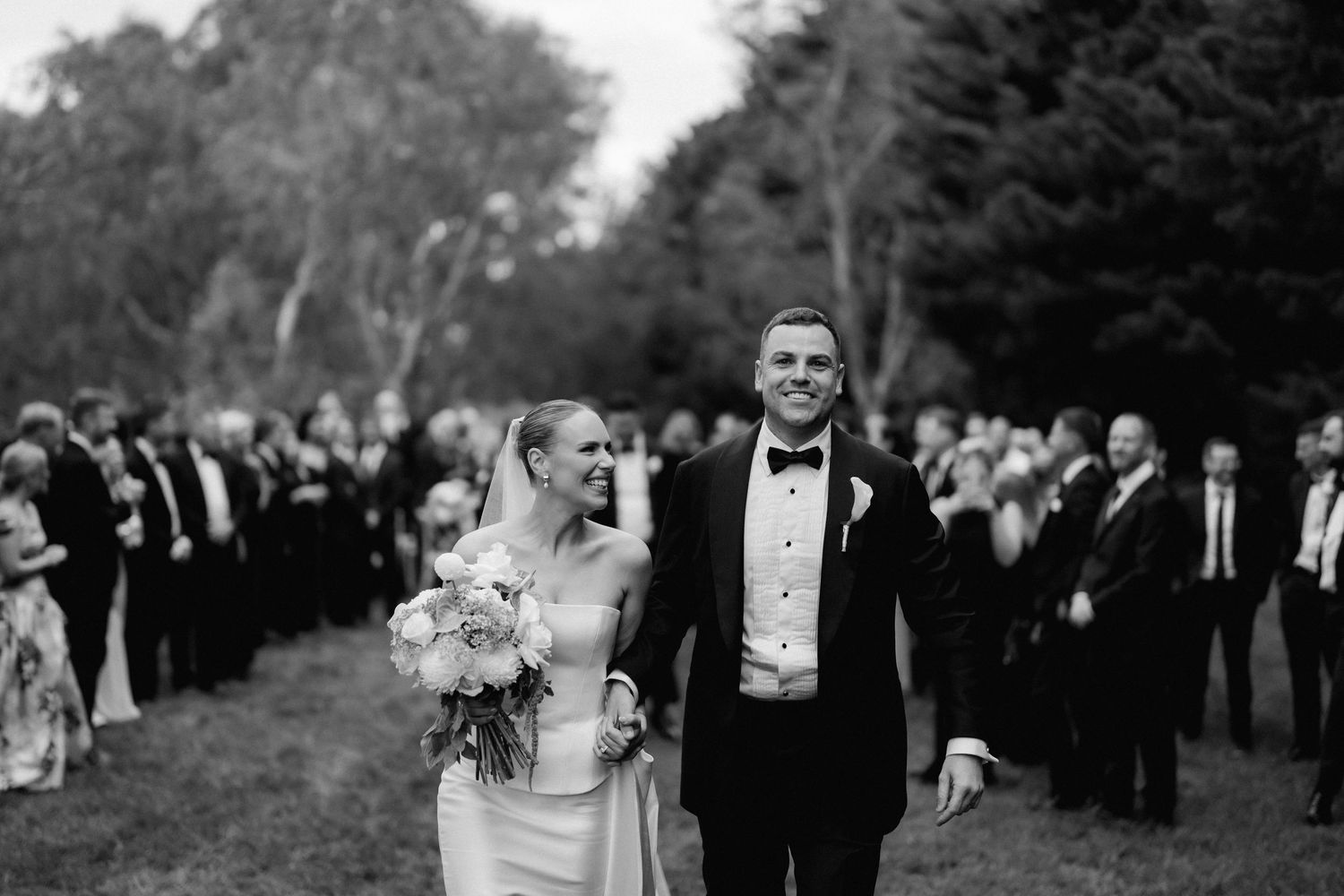 Black and white wedding photo of newlyweds walking past guests during an outdoor ceremony.