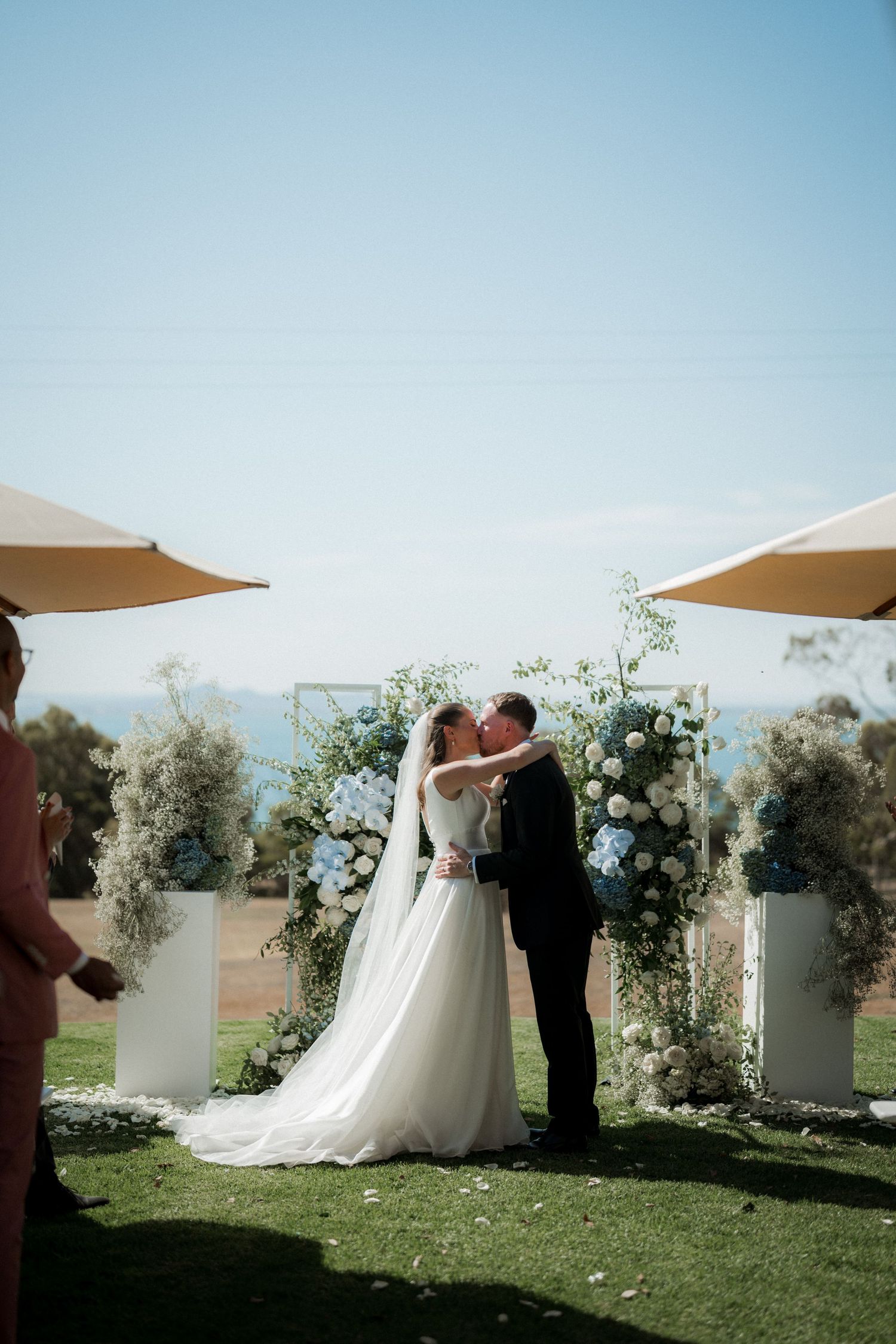 Outdoor wedding ceremony with couple kissing under a floral arch with beige umbrellas and mountain views.