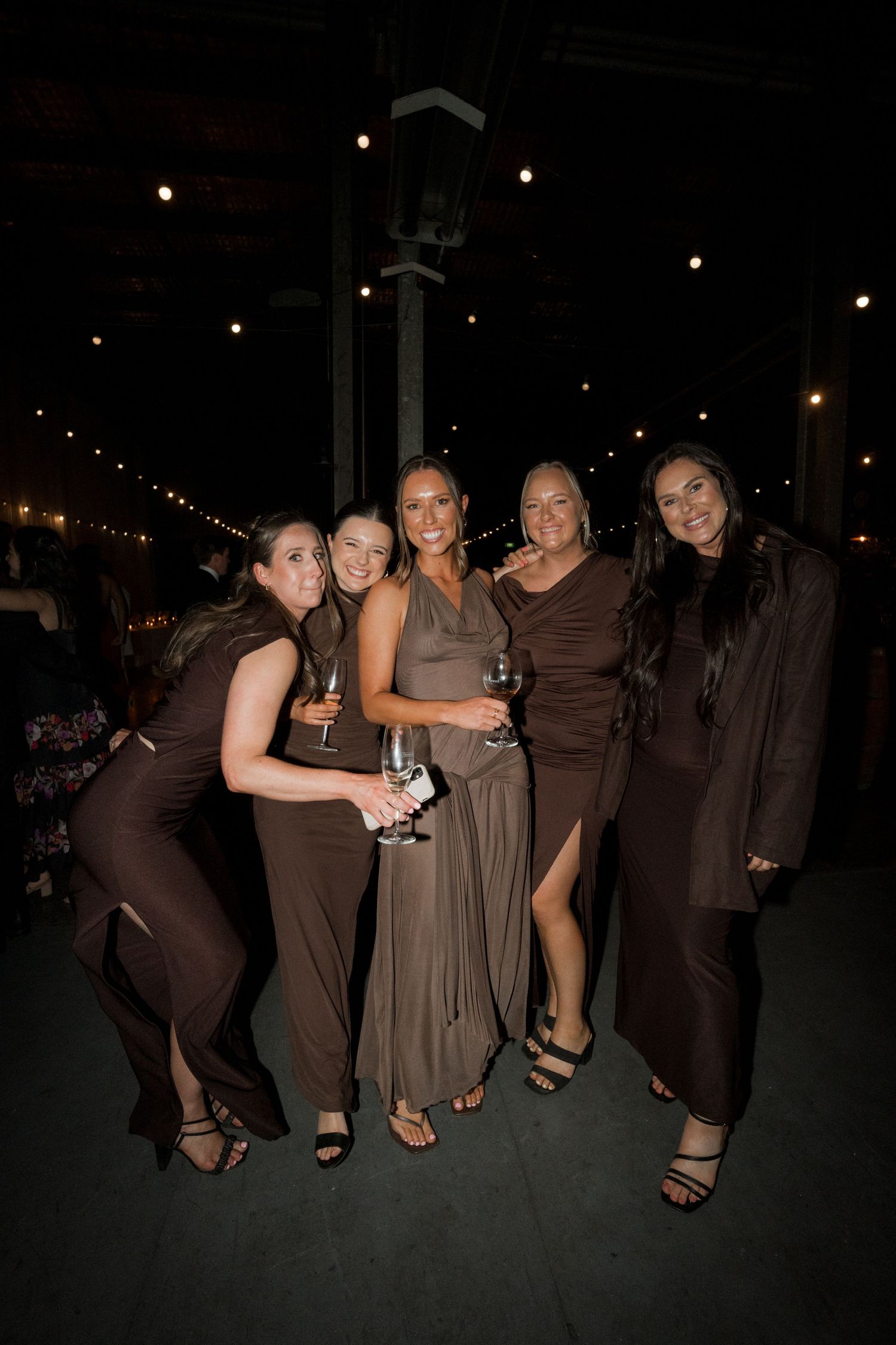 Group of friends in coordinating brown dresses pose together at an evening event.