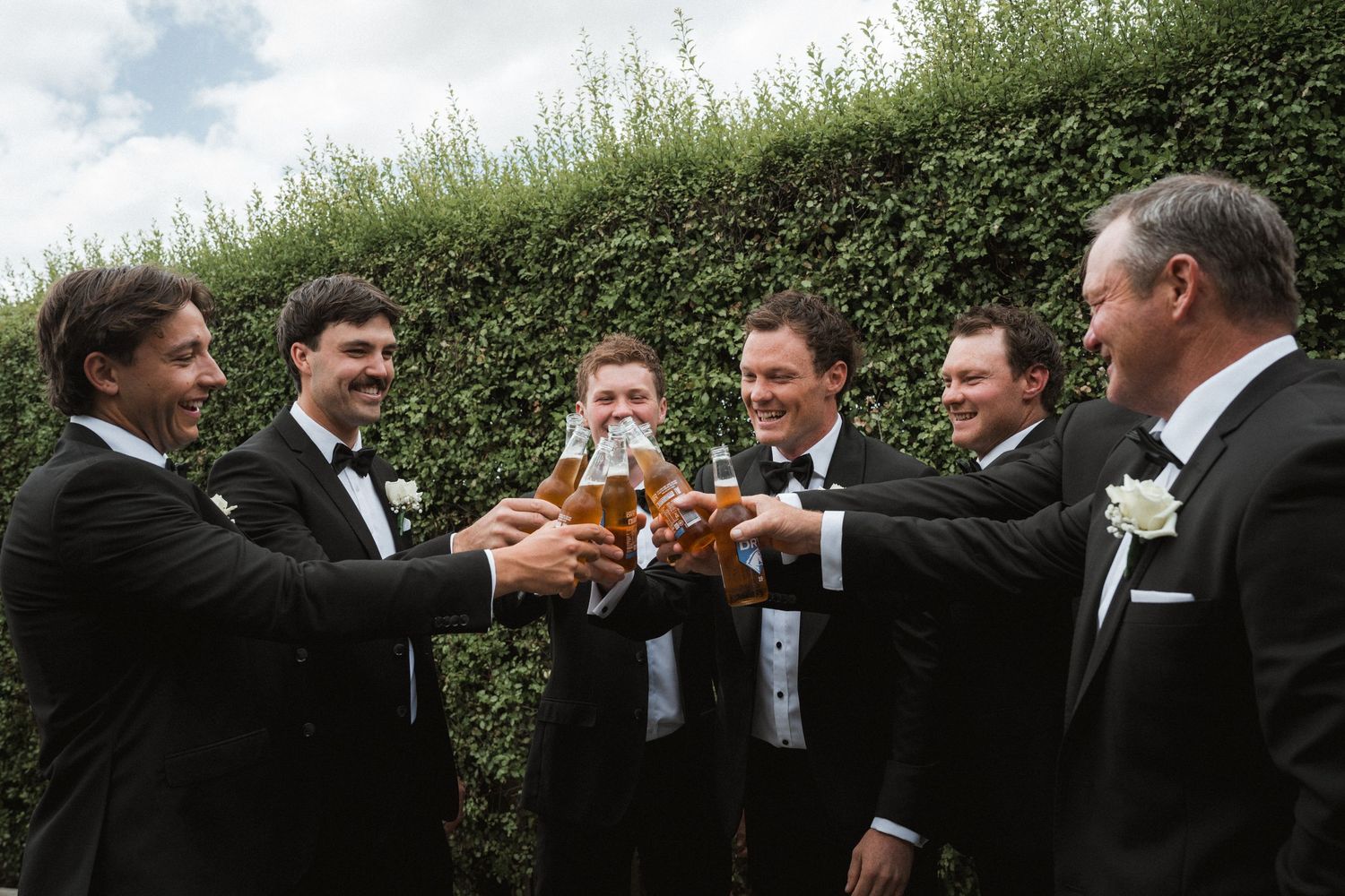 Groomsmen in formal attire toasting with beer glasses against a green hedge backdrop.