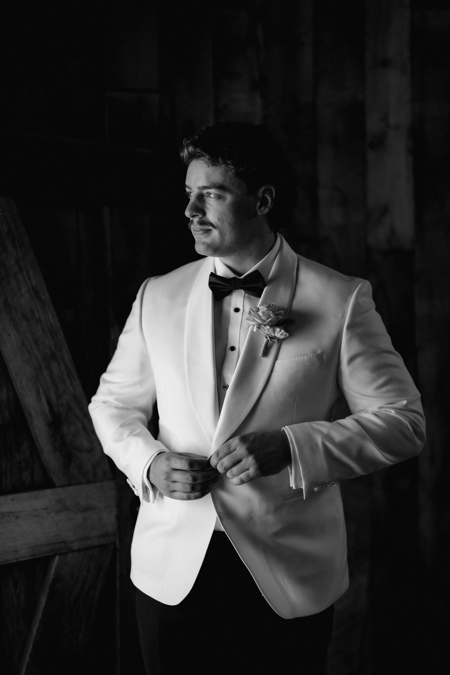 Groom in white tuxedo jacket and black bow tie adjusting his boutonniere in a black and white wedding photo.