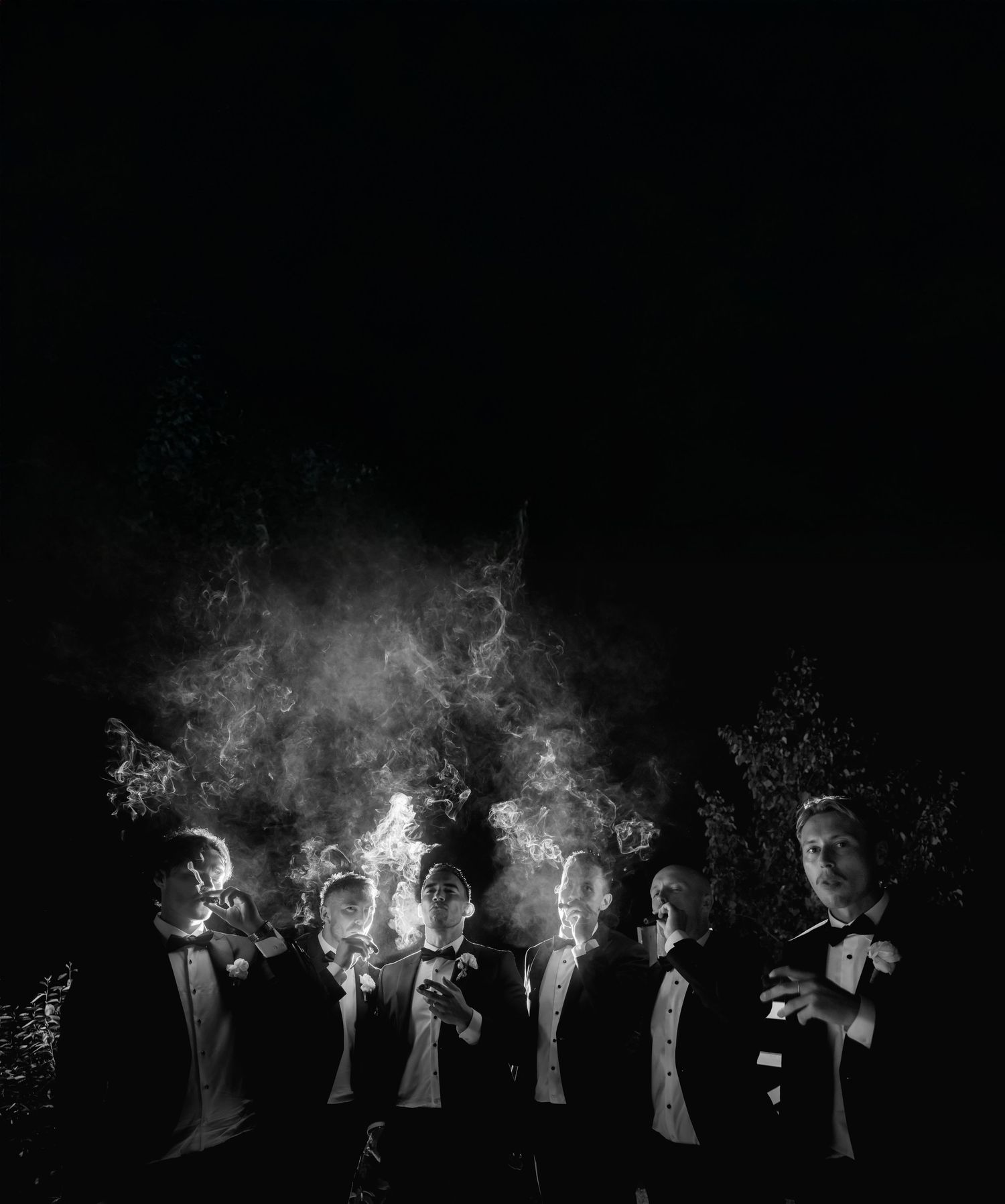 Black and white photo of six men in tuxedos smoking cigars at night with dramatic smoke clouds rising above them.
