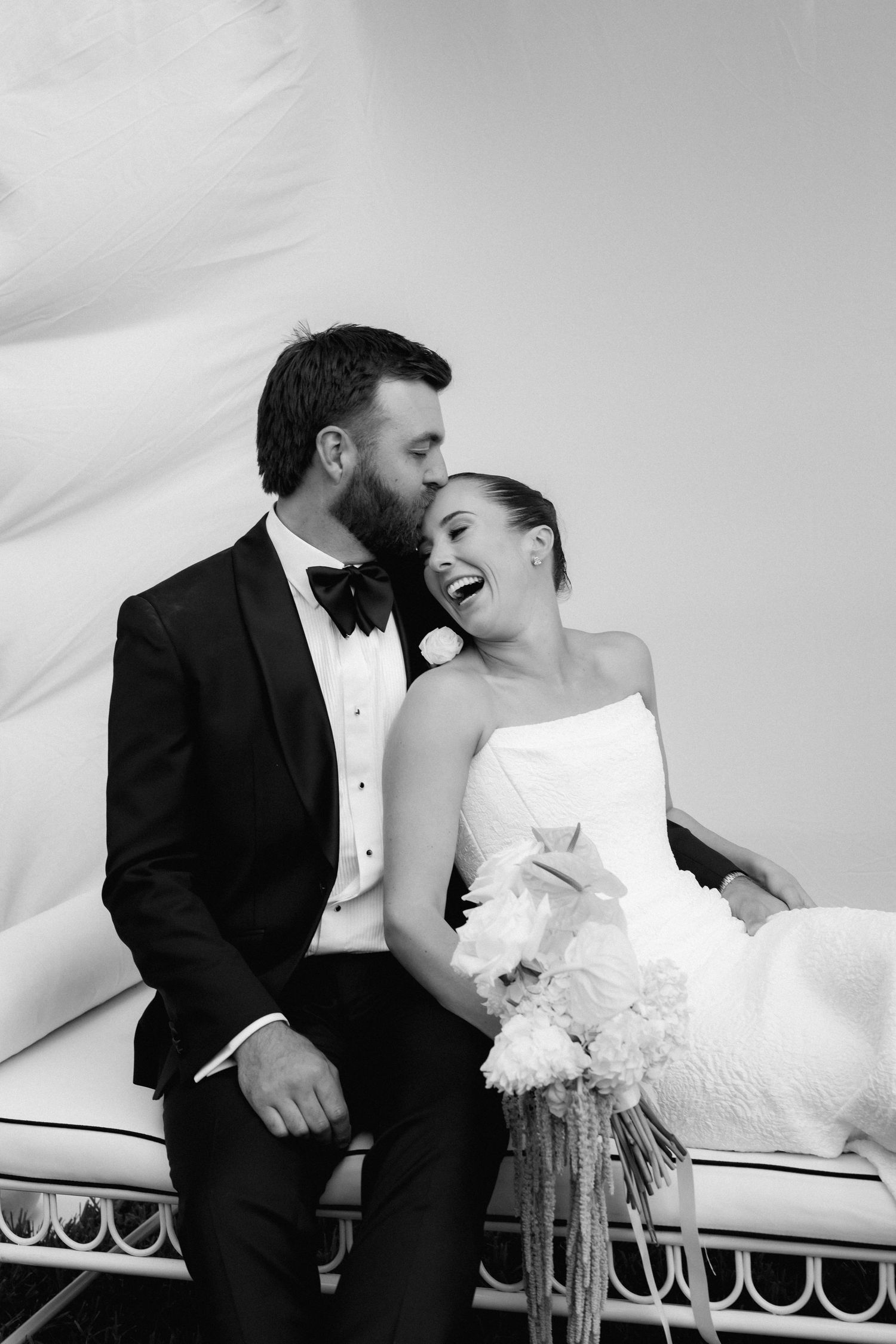 Bride and groom share a joyful moment seated together, with groom whispering in brides ear, black and white photo.