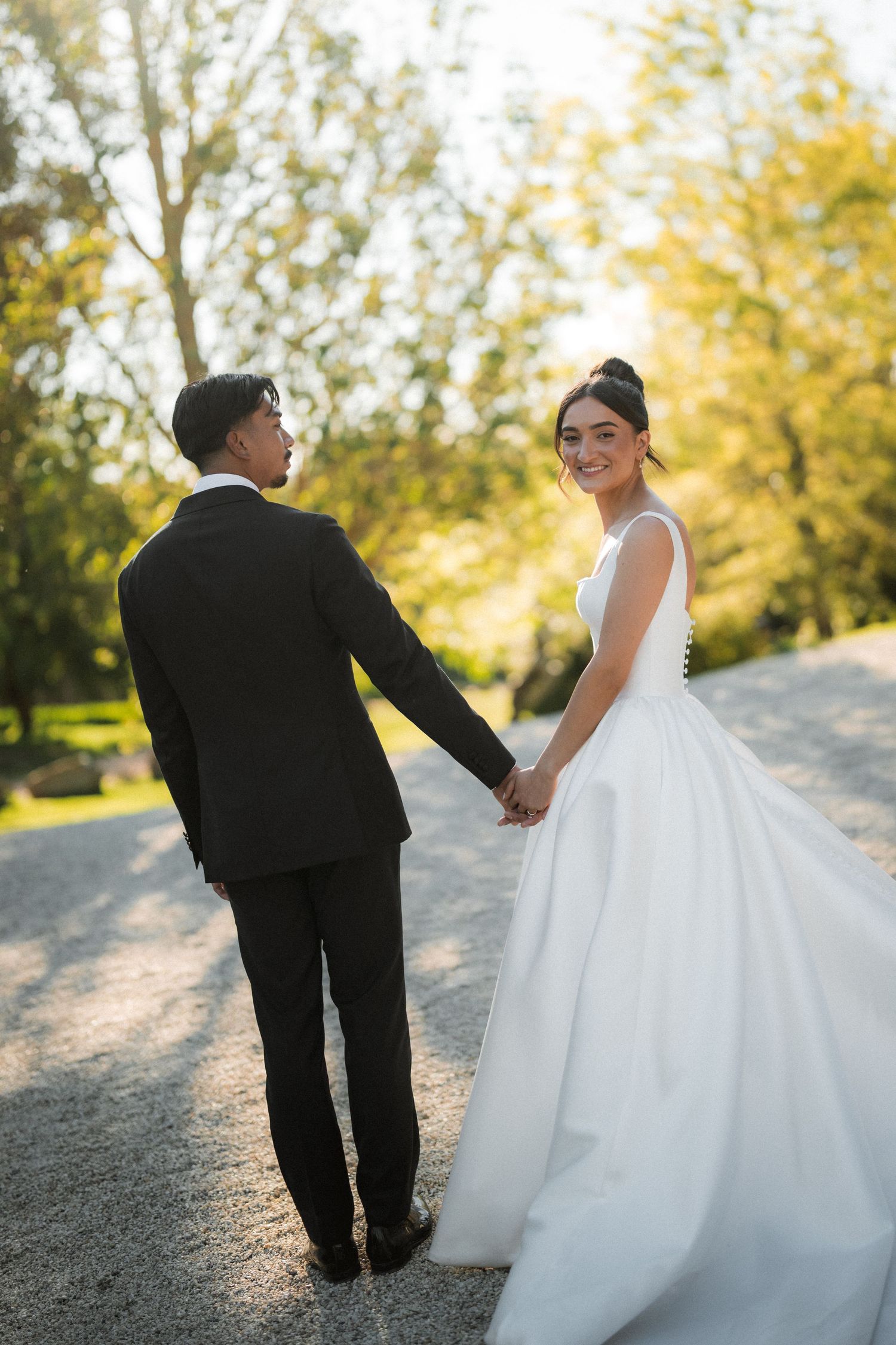 Bride in a white satin ball gown and groom in a dark suit hold hands on a gravel path surrounded by sunlit autumn trees.