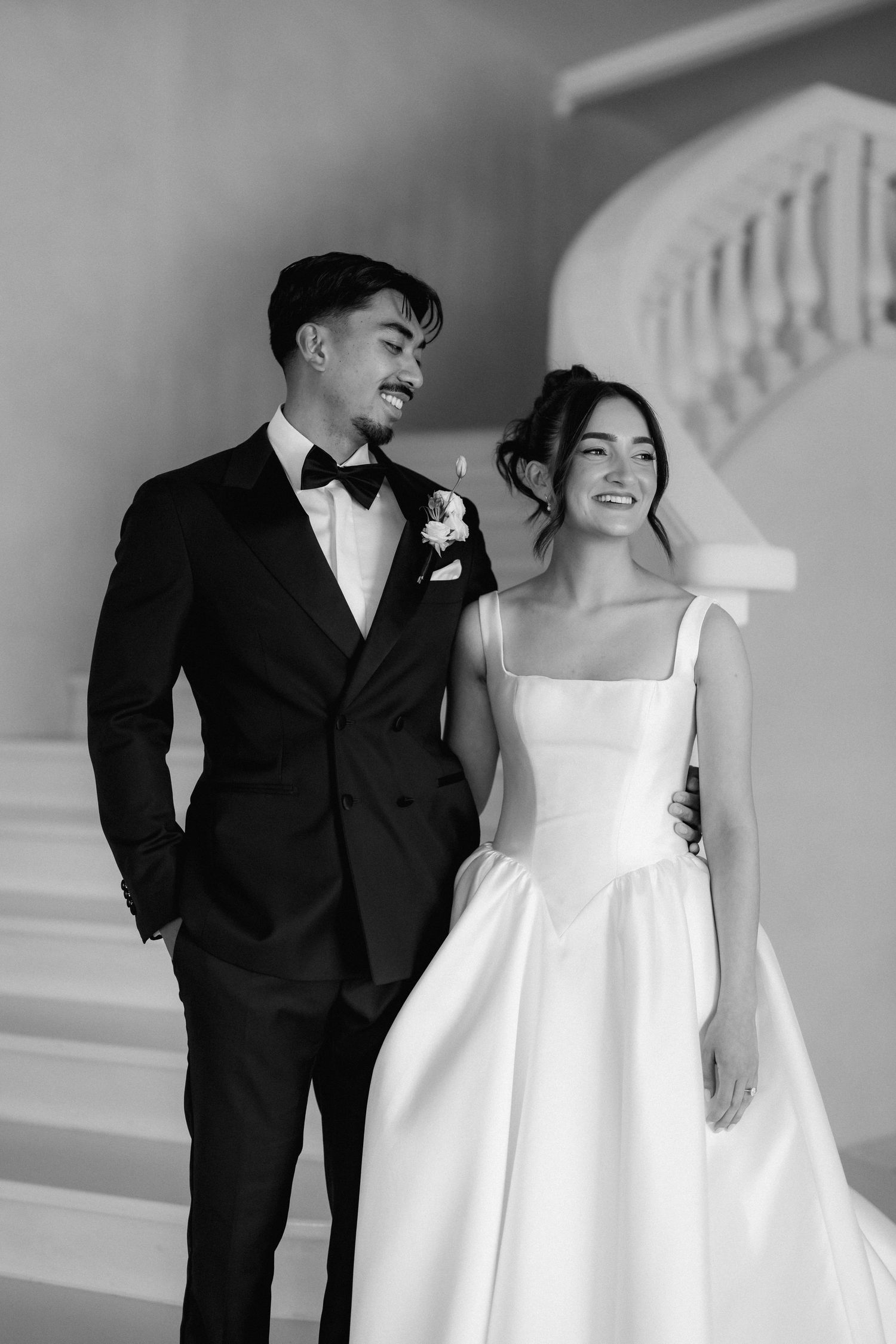 Black and white wedding portrait of a smiling groom in a tuxedo and bow tie beside a bride in a satin gown on a grand staircase.
