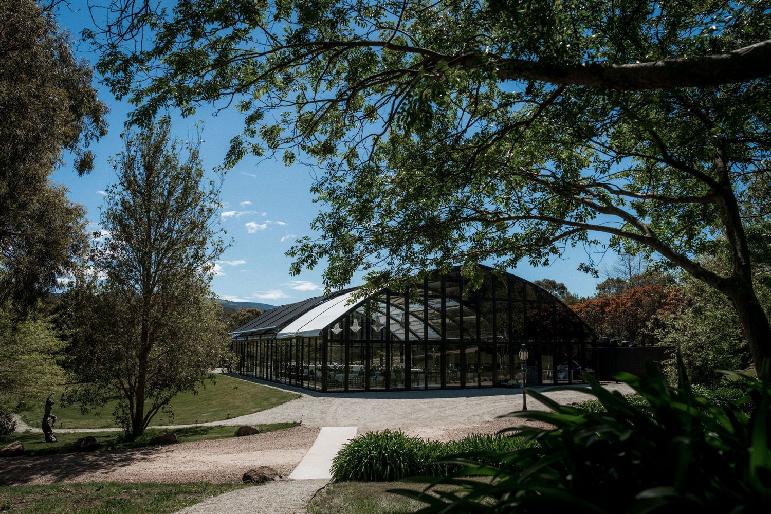 Glass greenhouse conservatory surrounded by lush trees and garden paths under a blue sky with scattered clouds.