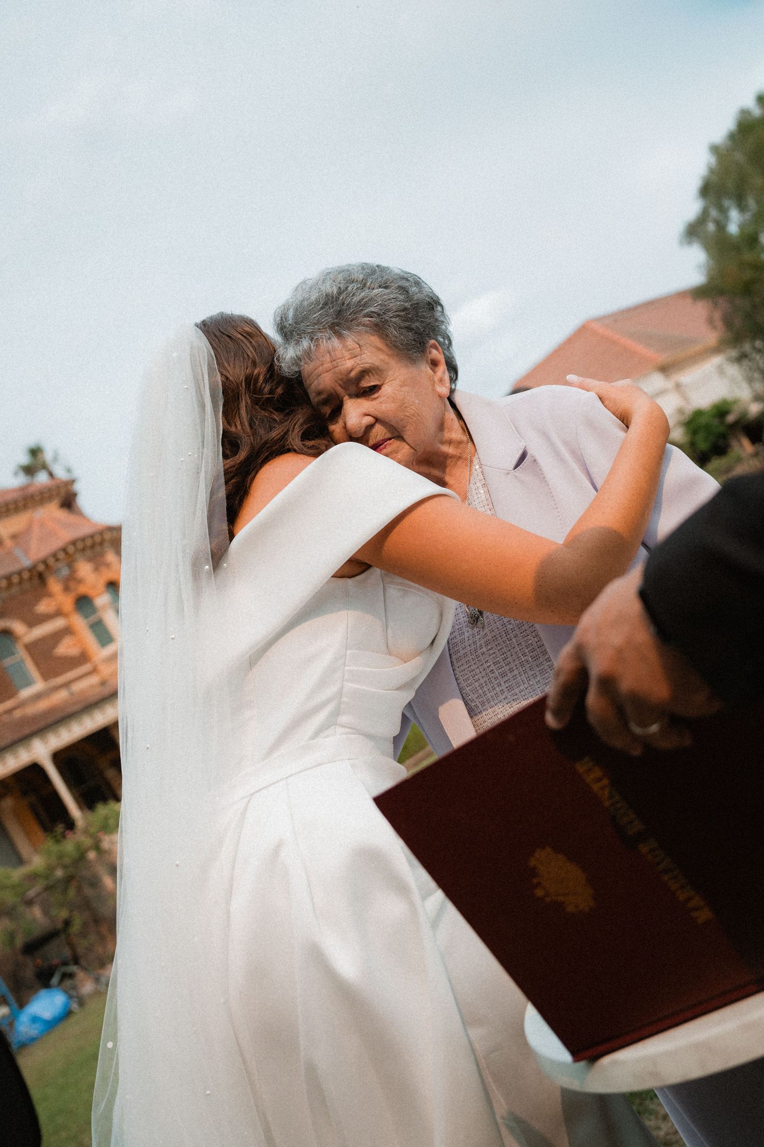 A bride in a white dress shares an emotional embrace during an outdoor wedding ceremony.
