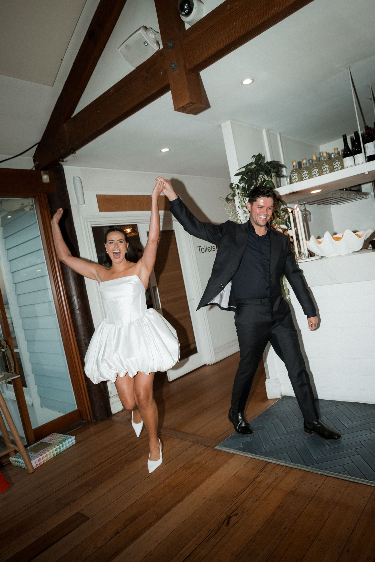 Two wedding guests joyfully dance and leap together in a rustic room with wooden beams and hardwood floors.