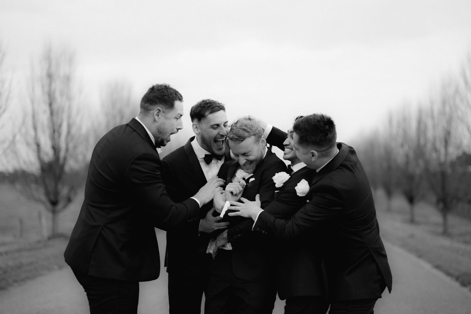 Groomsmen in black tuxedos share a lighthearted moment during wedding celebrations.