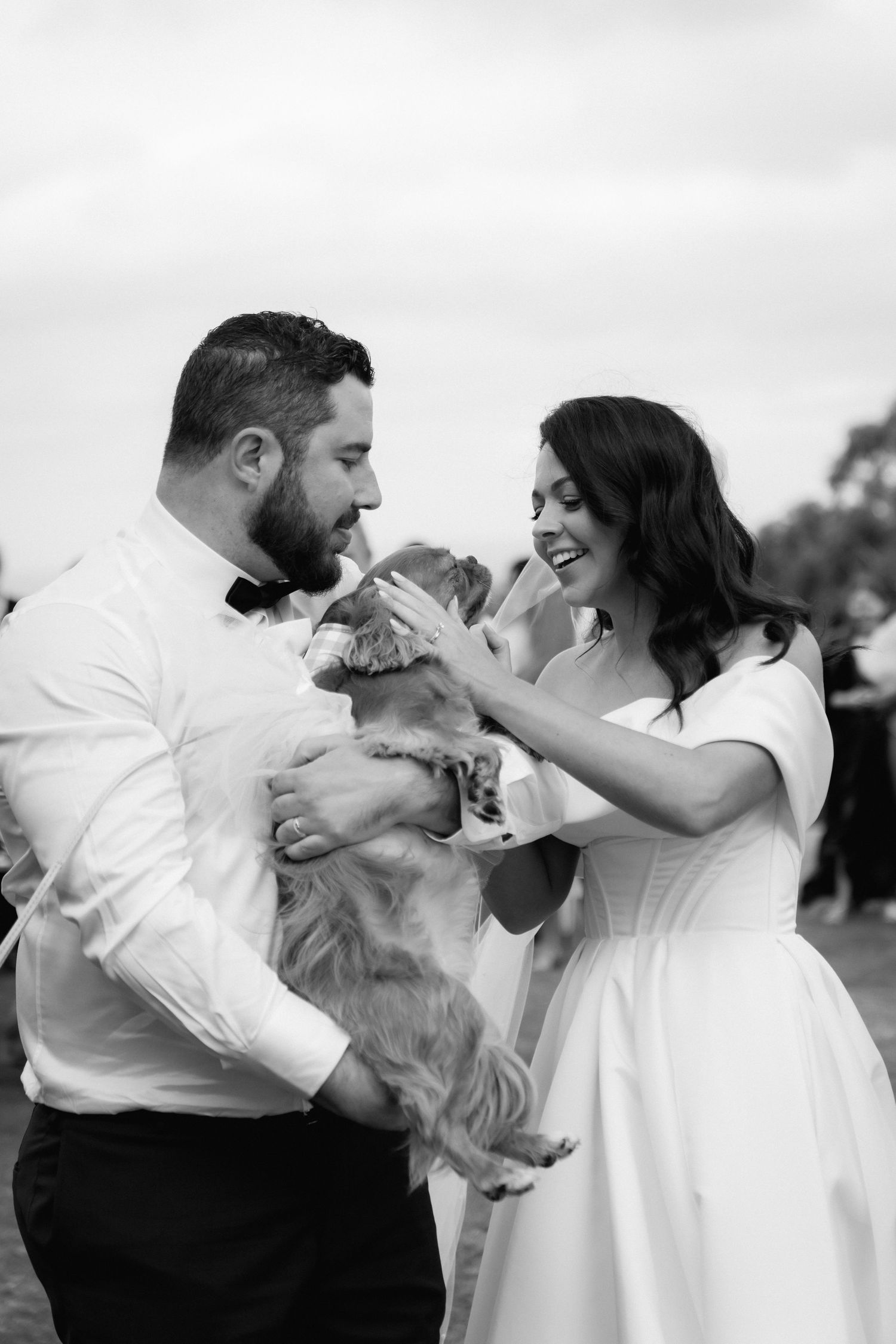 A couple in wedding attire share a joyful moment with their fluffy dog during an outdoor photoshoot.