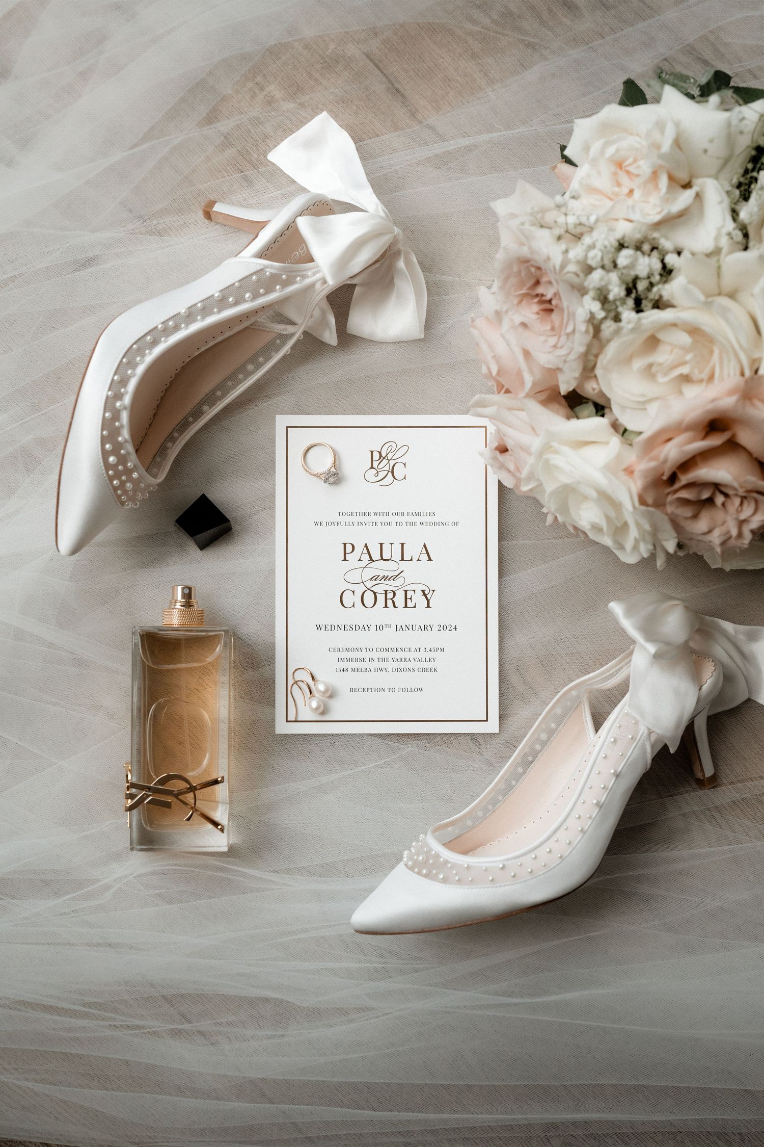Elegant wedding details including white bridal shoes, floral invitation, and perfume bottle on tulle fabric.