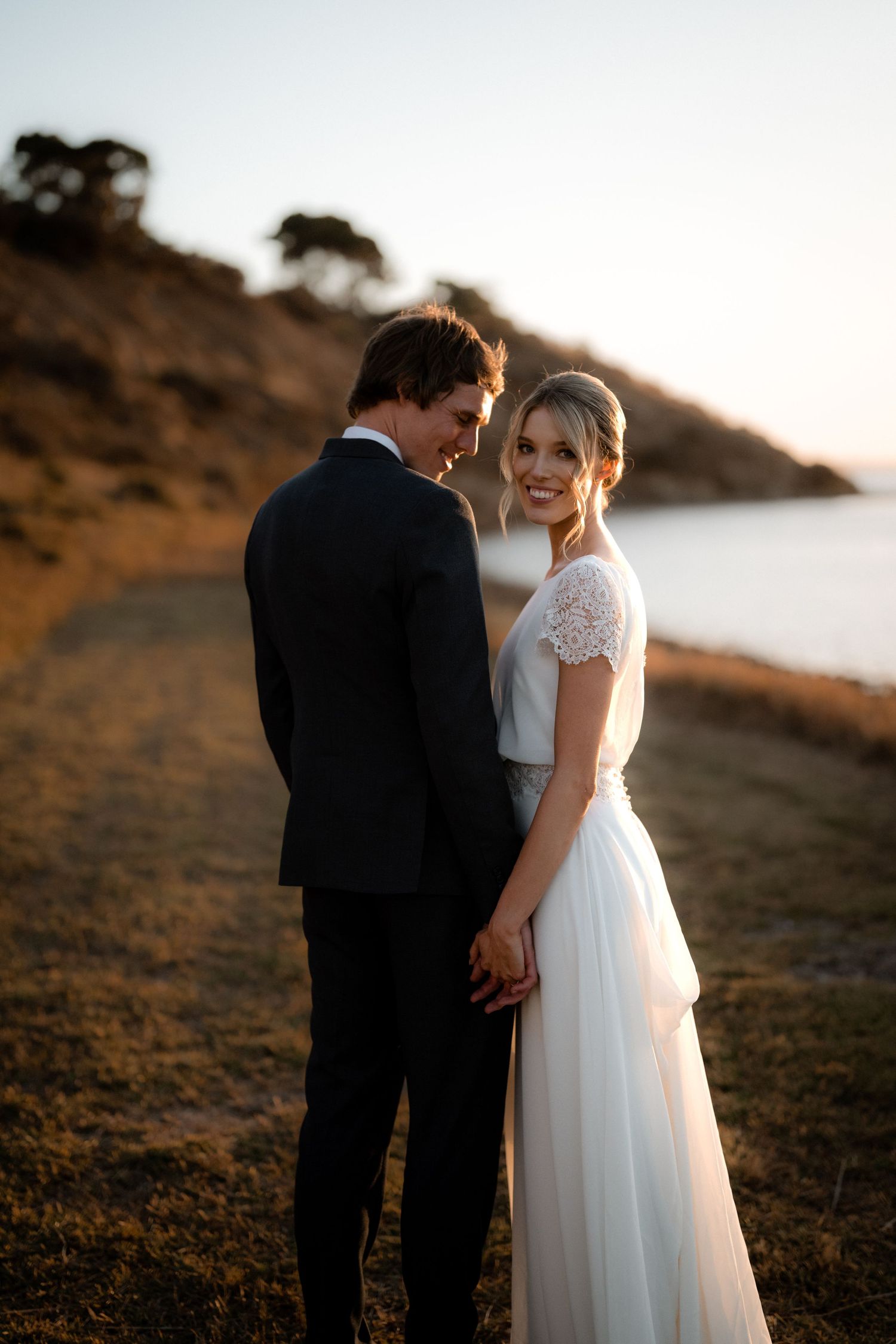A couple in traditional wedding attire share an intimate moment at sunset on a coastal hillside.