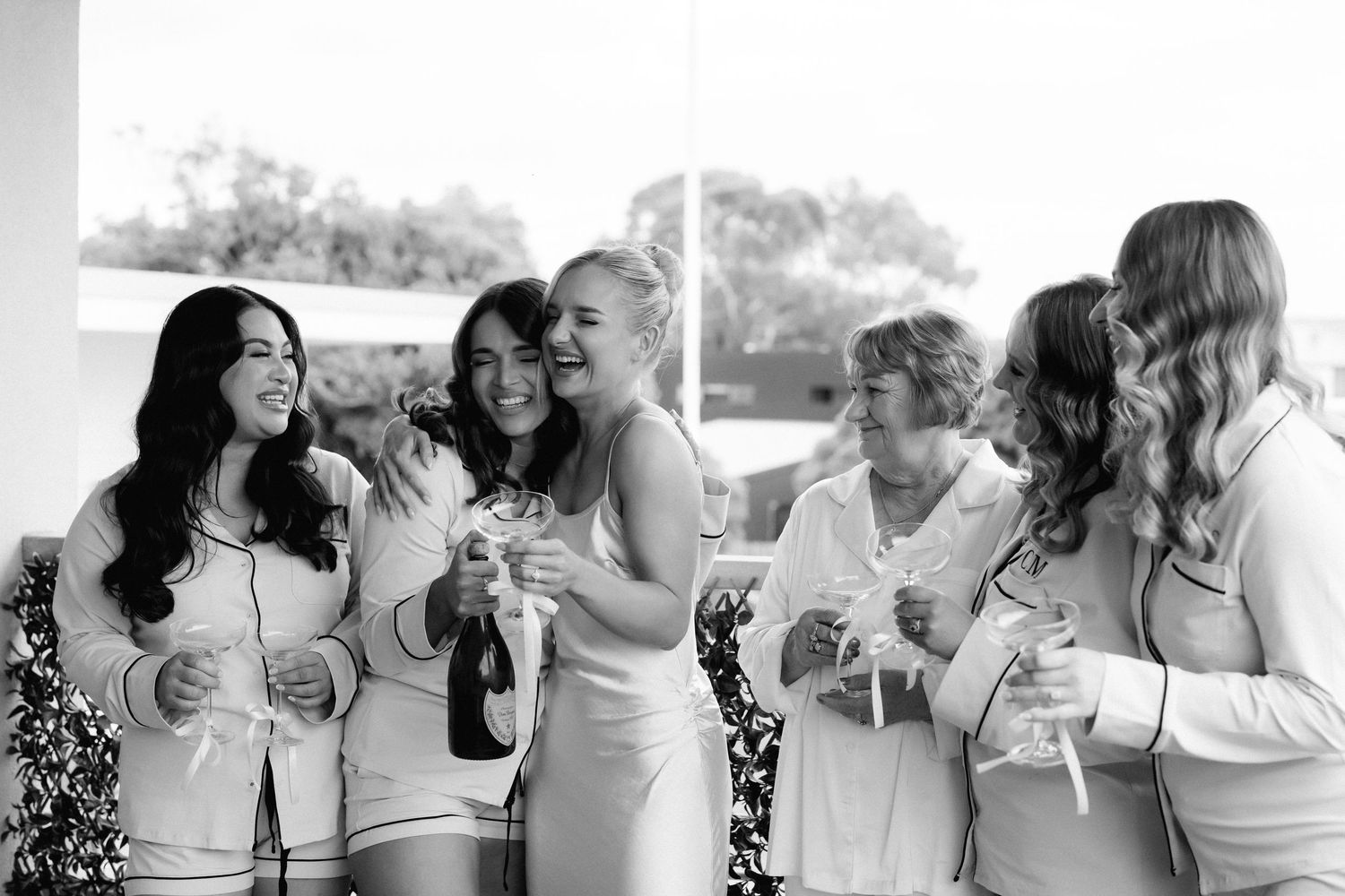 A group of wedding guests in light colored dresses share joyful laughter while holding drinks outdoors.