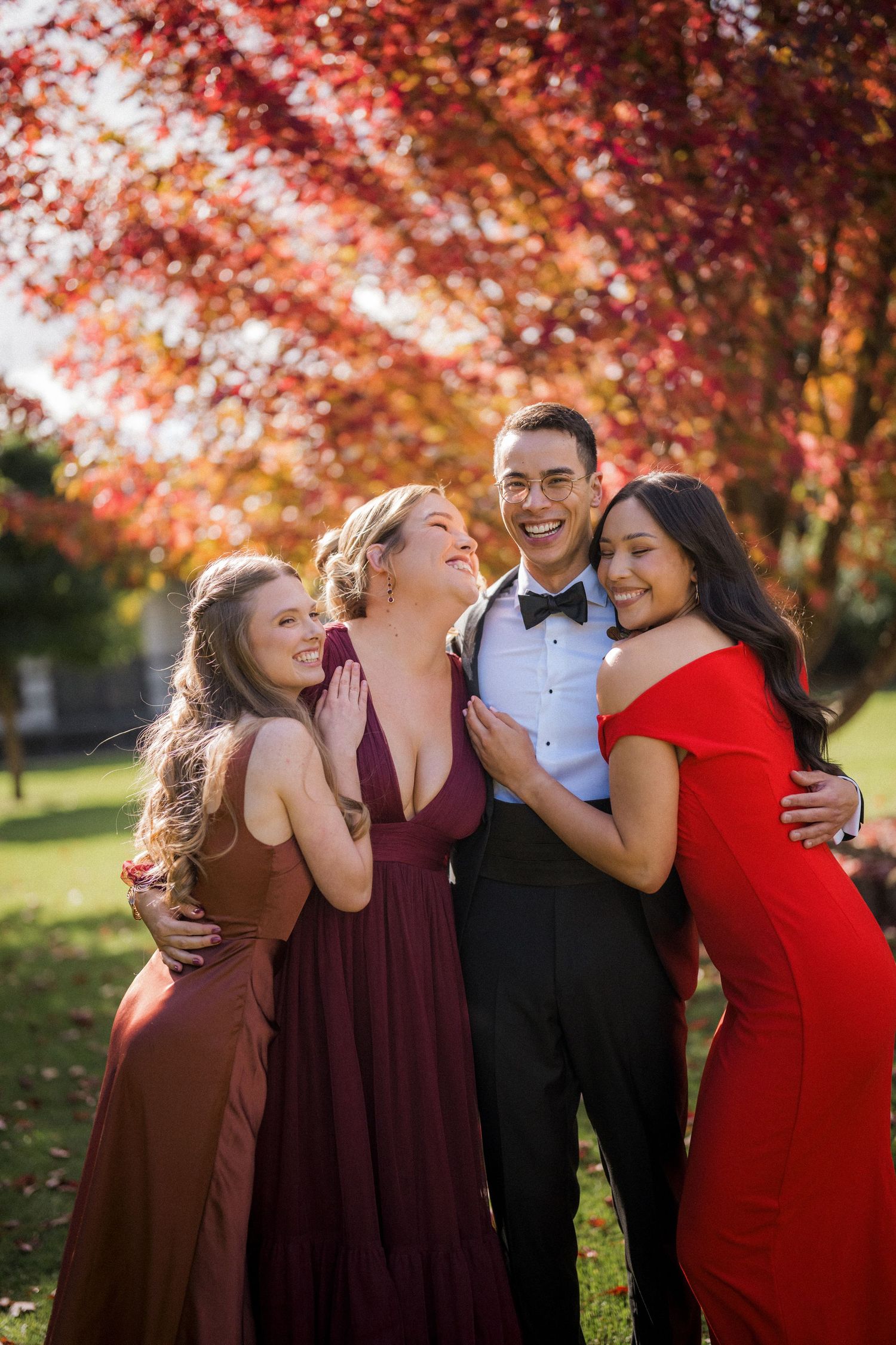 Four elegantly dressed friends pose together outdoors, laughing joyfully beneath vibrant red autumn foliage.