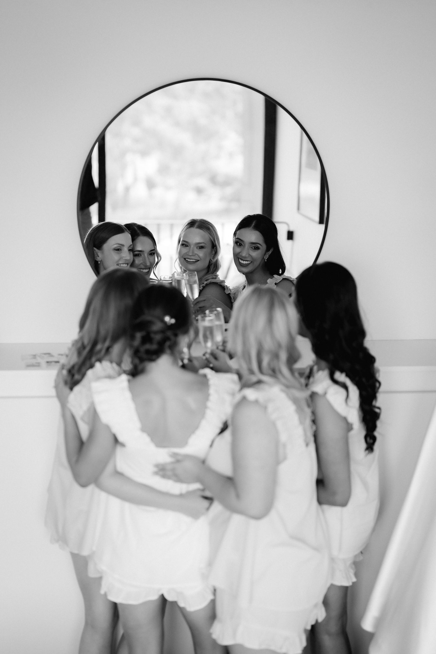 Black and white photo of attendants in white dresses gathered around a mirror during pre-wedding preparations.