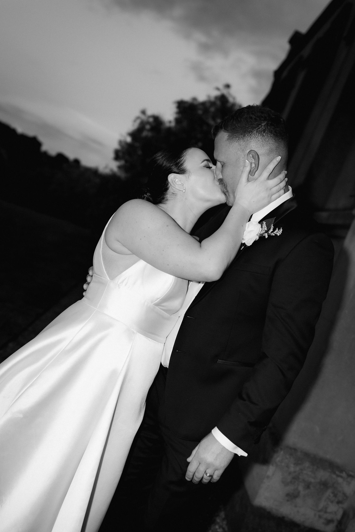 A romantic black and white portrait of a couple sharing an intimate kiss at sunset against moody skies.