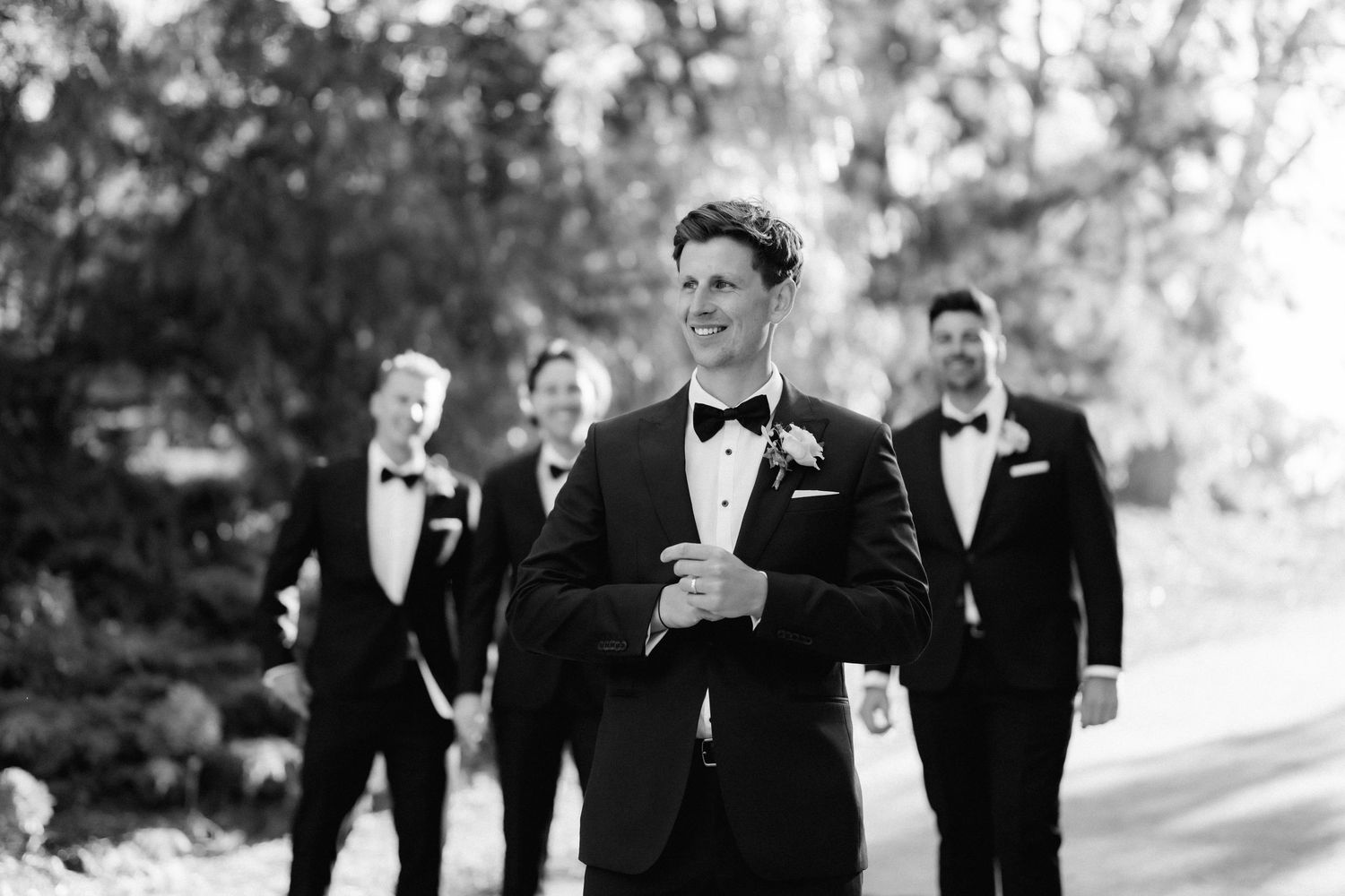 Groomsmen in black tuxedos walking together outdoors at a formal wedding.