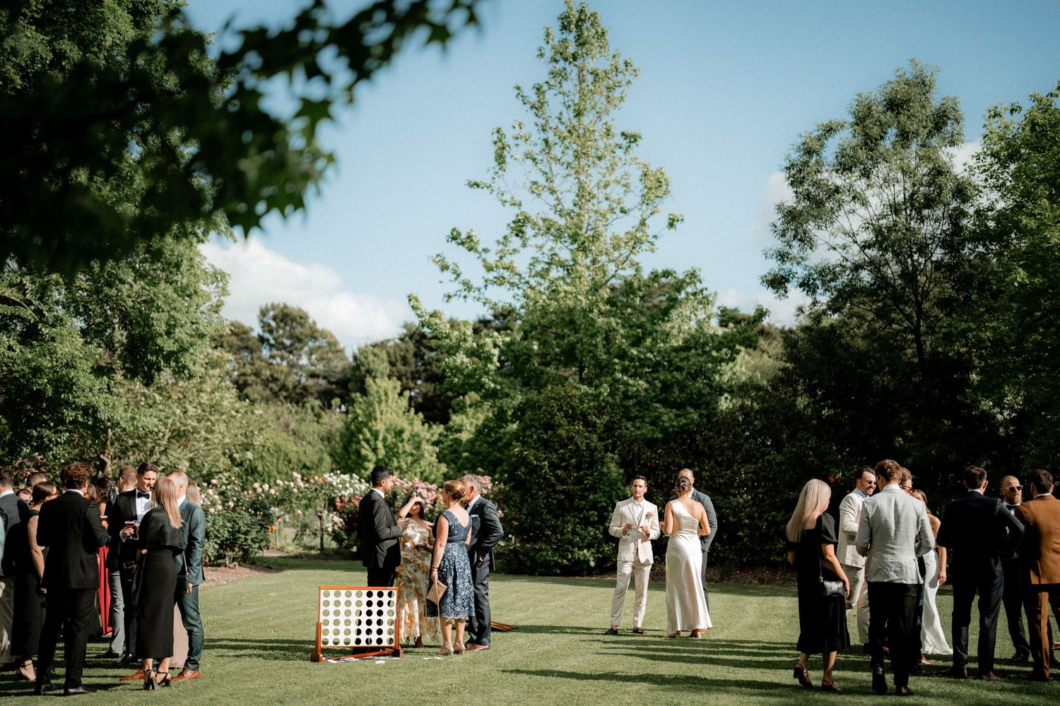 Wedding guests gather outside on a sunny day surrounded by green trees and lawn during an outdoor ceremony.