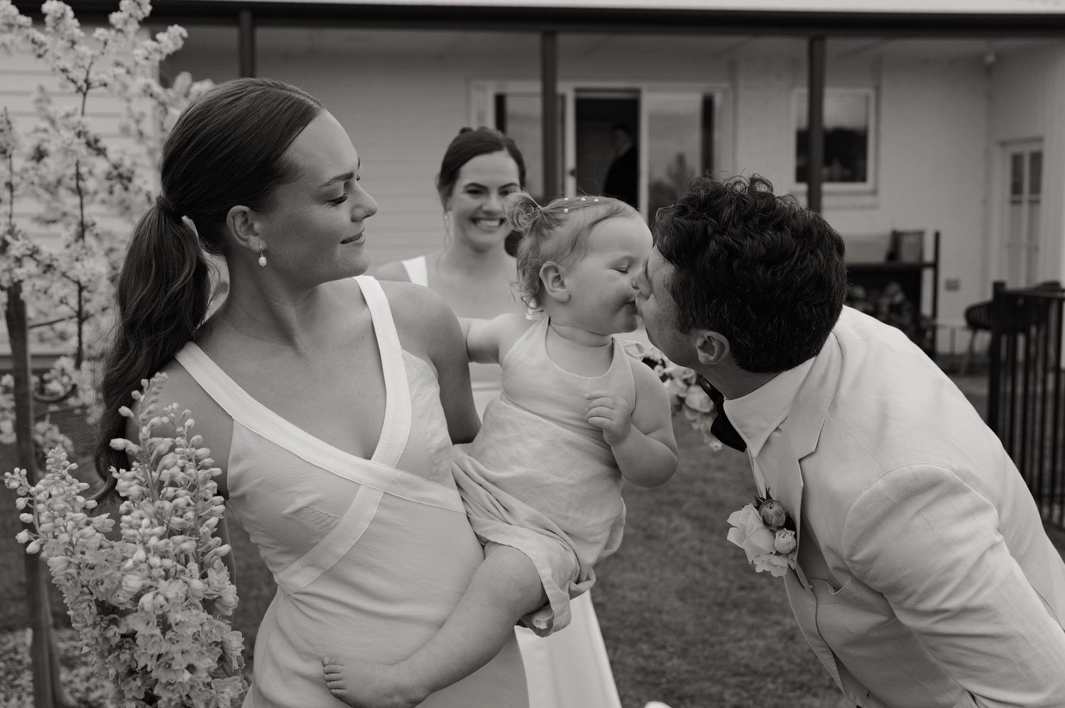 Black and white wedding photo of a man kissing a baby held by a woman in a white dress outdoors.