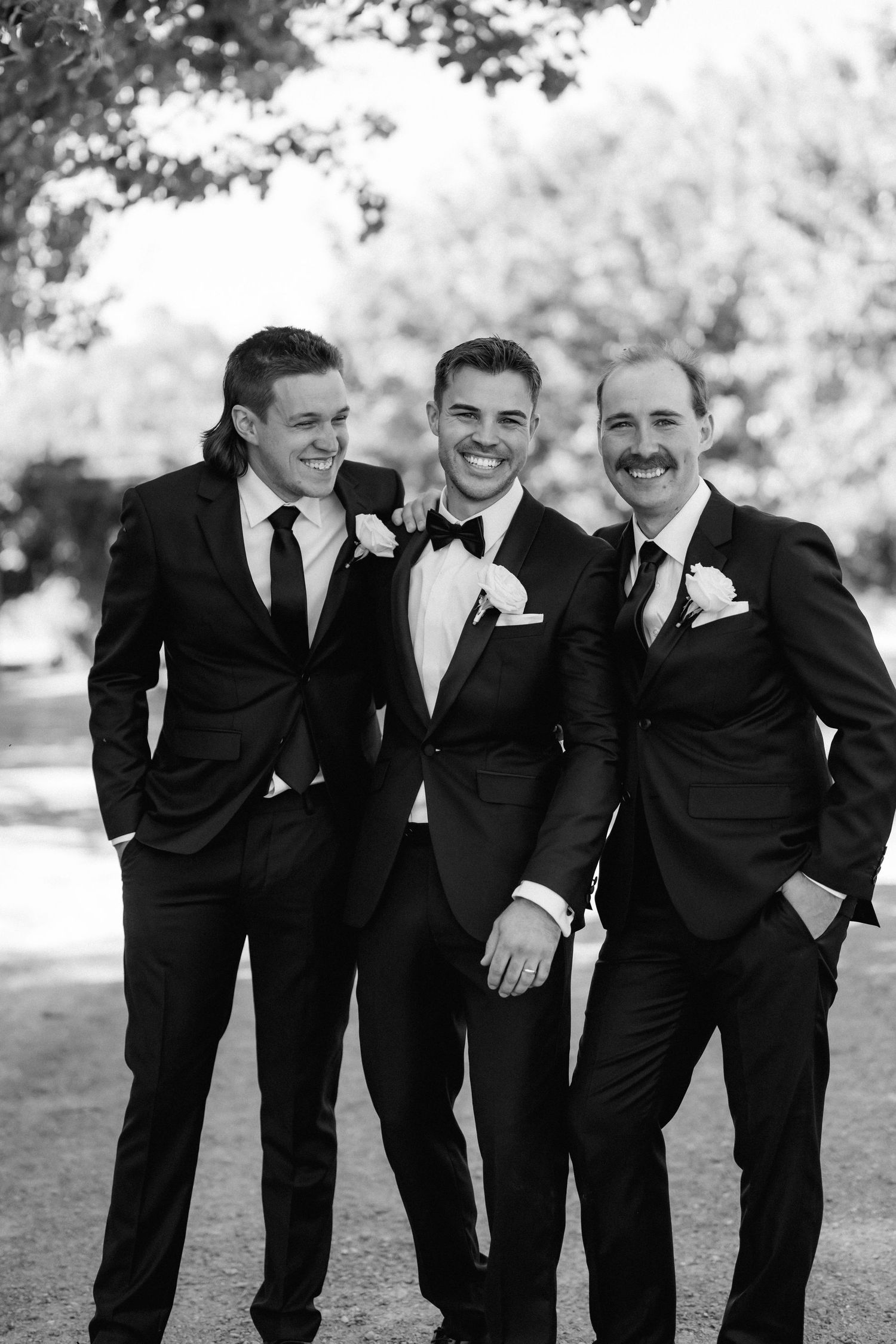 Groomsmen in black tuxedos share a candid laughing moment outdoors.