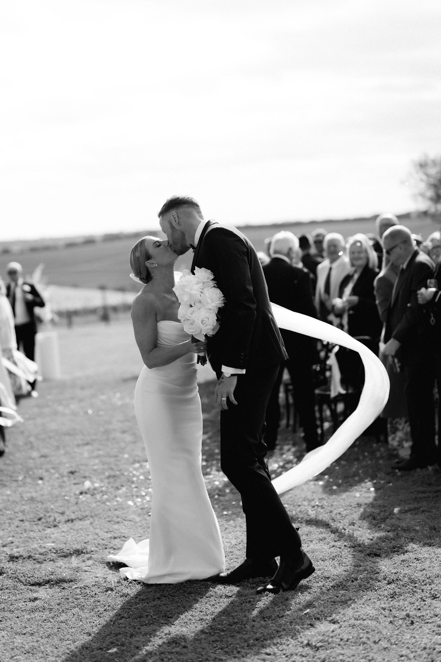 Wedding couple dances outdoors with guests watching in black and white photography.