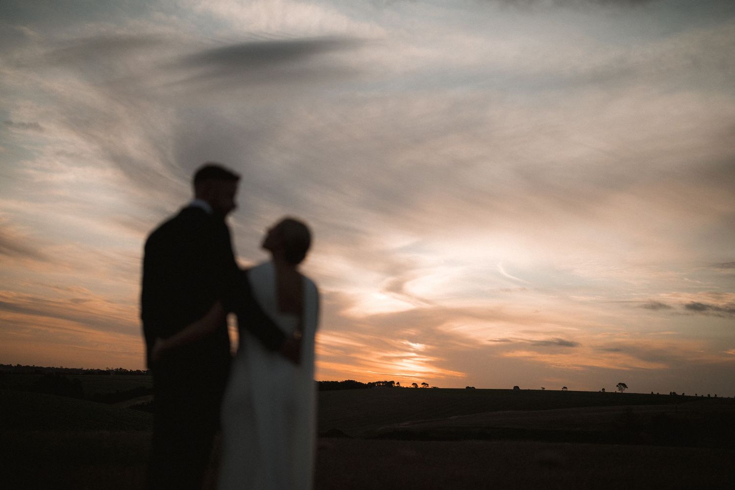 Silhouettes of two people stand together against a dramatic sunset sky with orange clouds.