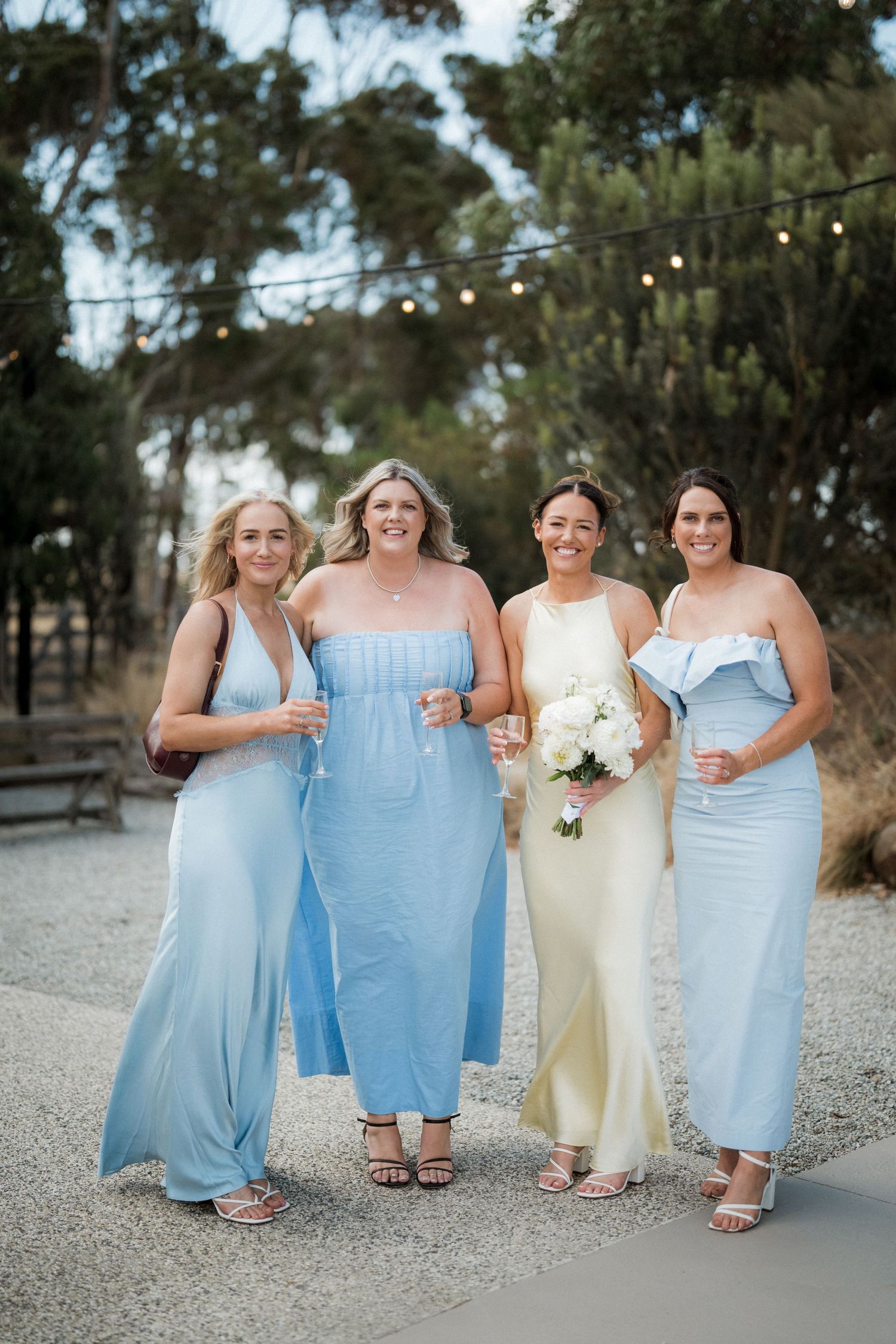A bridal party wearing light blue dresses and a cream gown pose together on a gravel path with string lights overhead.