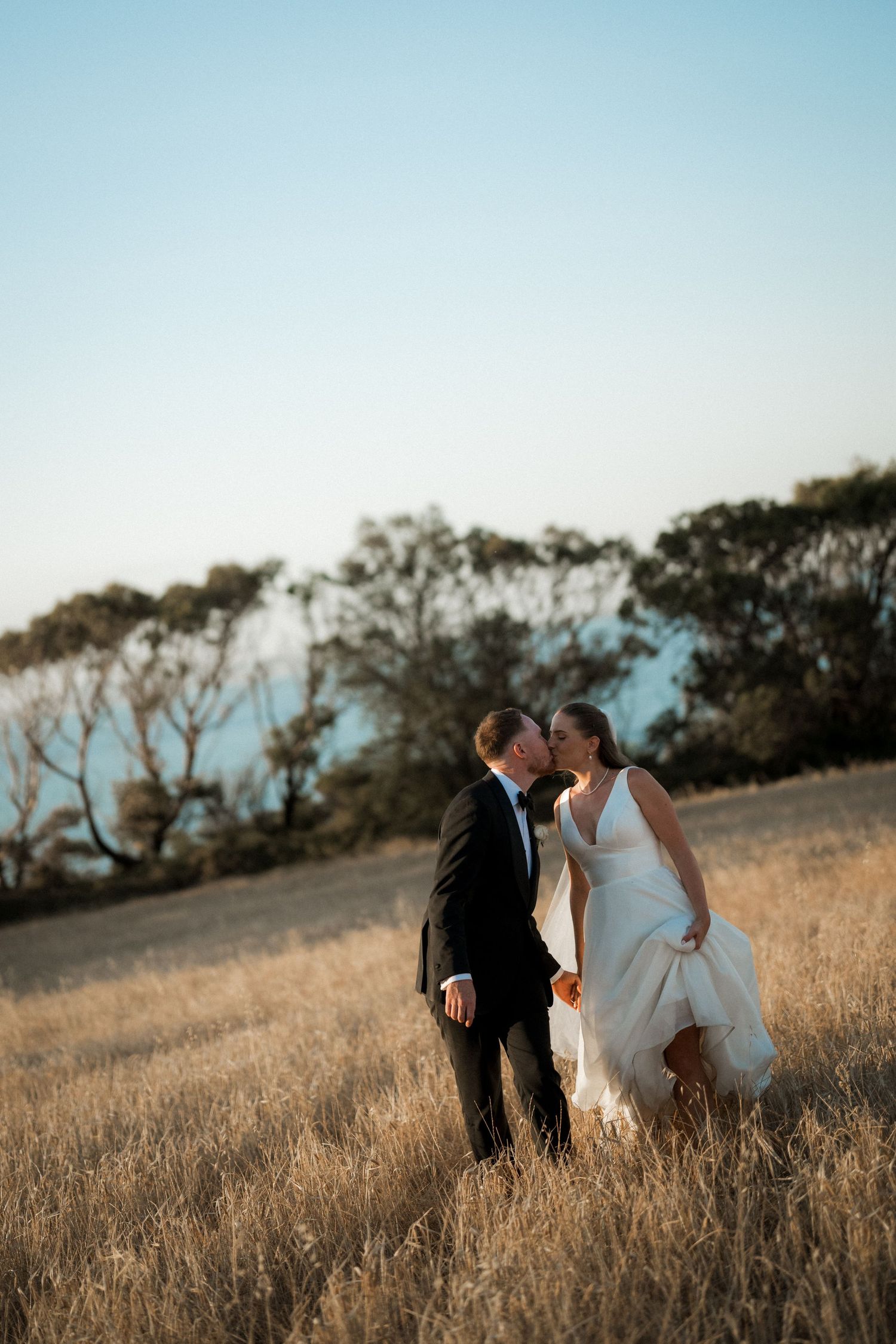 Couple in wedding attire walking through golden grass field with trees in background.