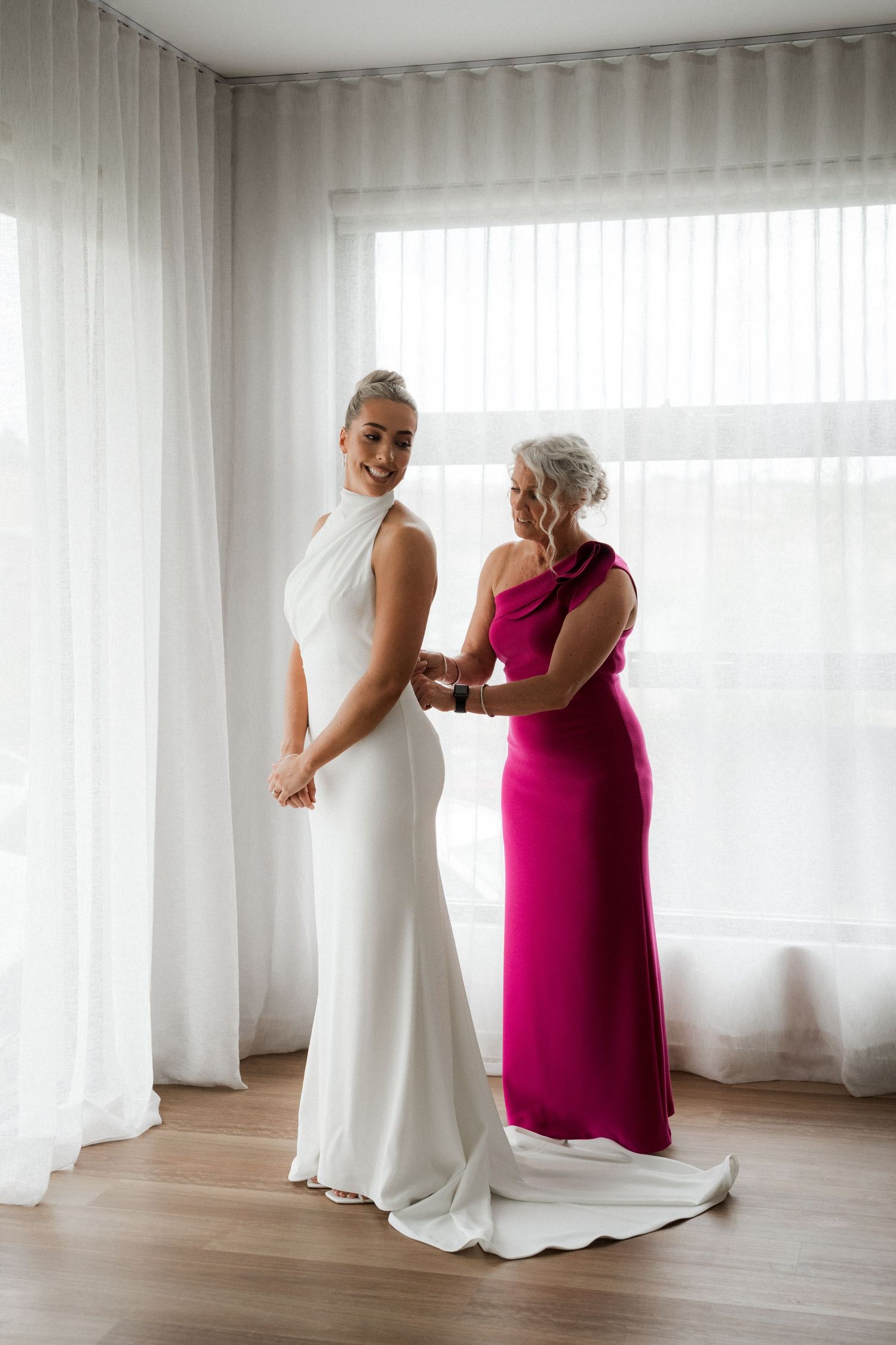 Two people sharing an intimate moment while adjusting a wedding dress near bright windows.